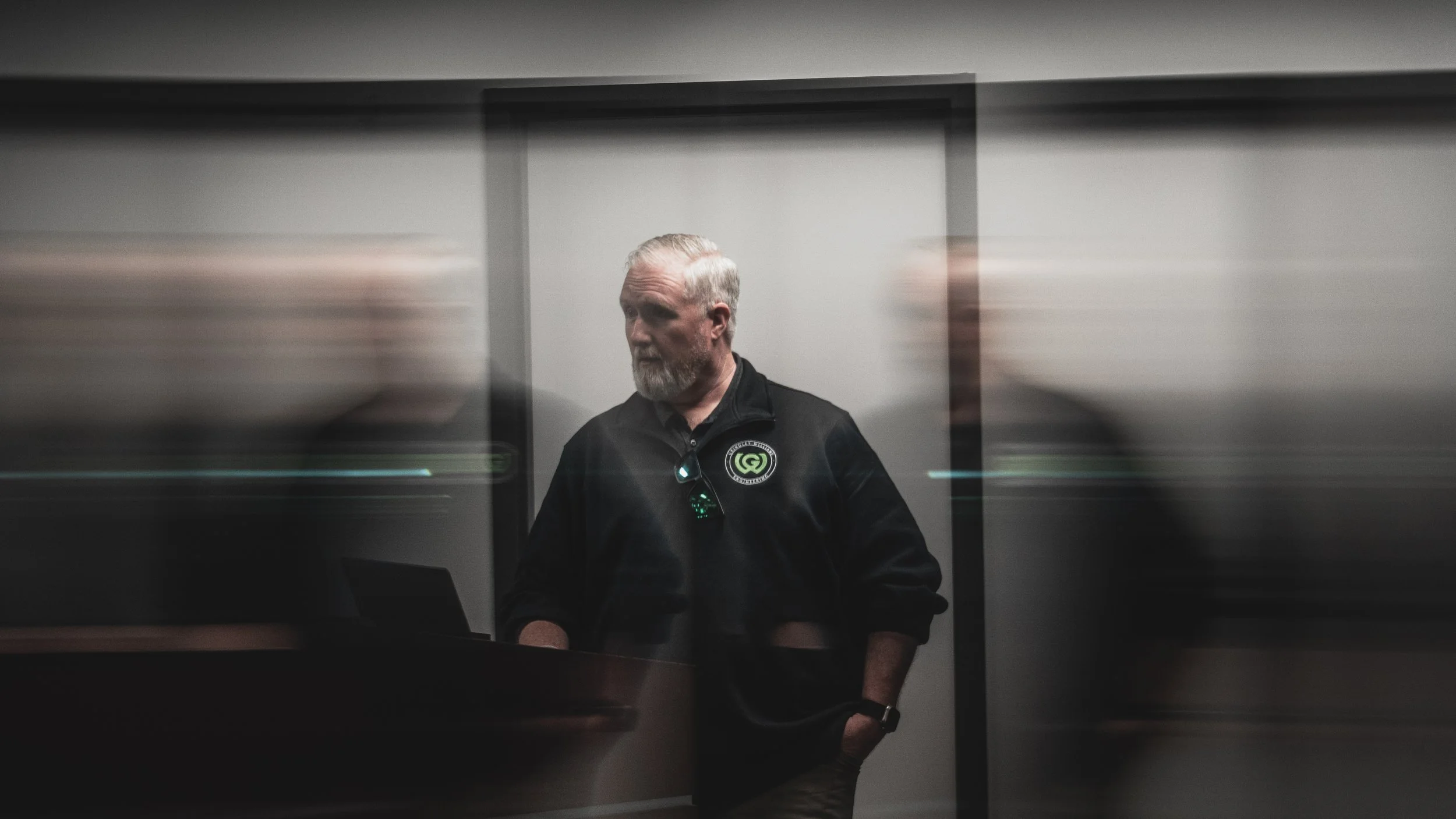 A man with gray hair and beard wearing a black jacket with a green and white patch stands in front of a plain wall, with blurred figures on either side.