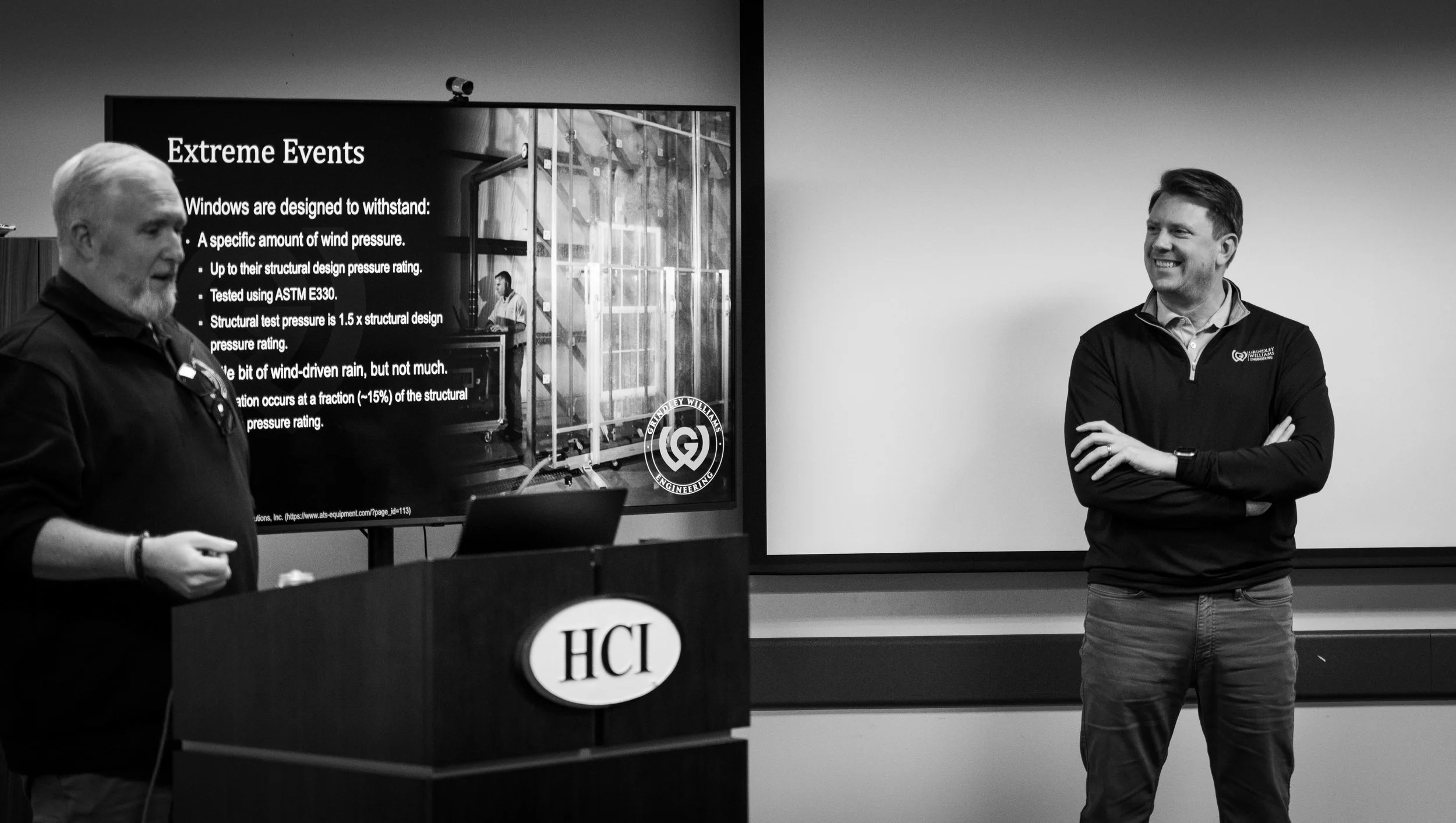 Two men giving a presentation in a conference room with a large screen displaying information about extreme events and structural testing.
