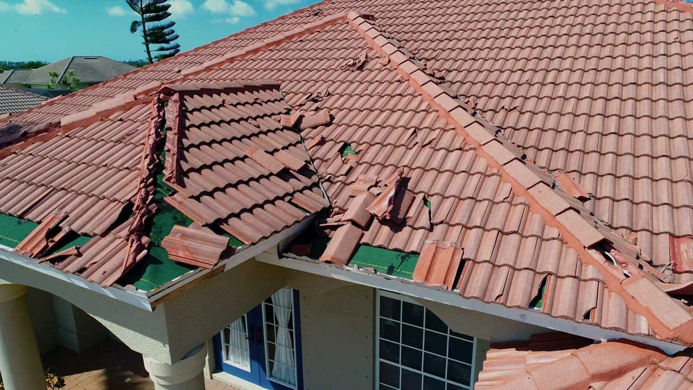 A house with a damaged red tile roof, with several missing or broken tiles, and a new section of green tiles.