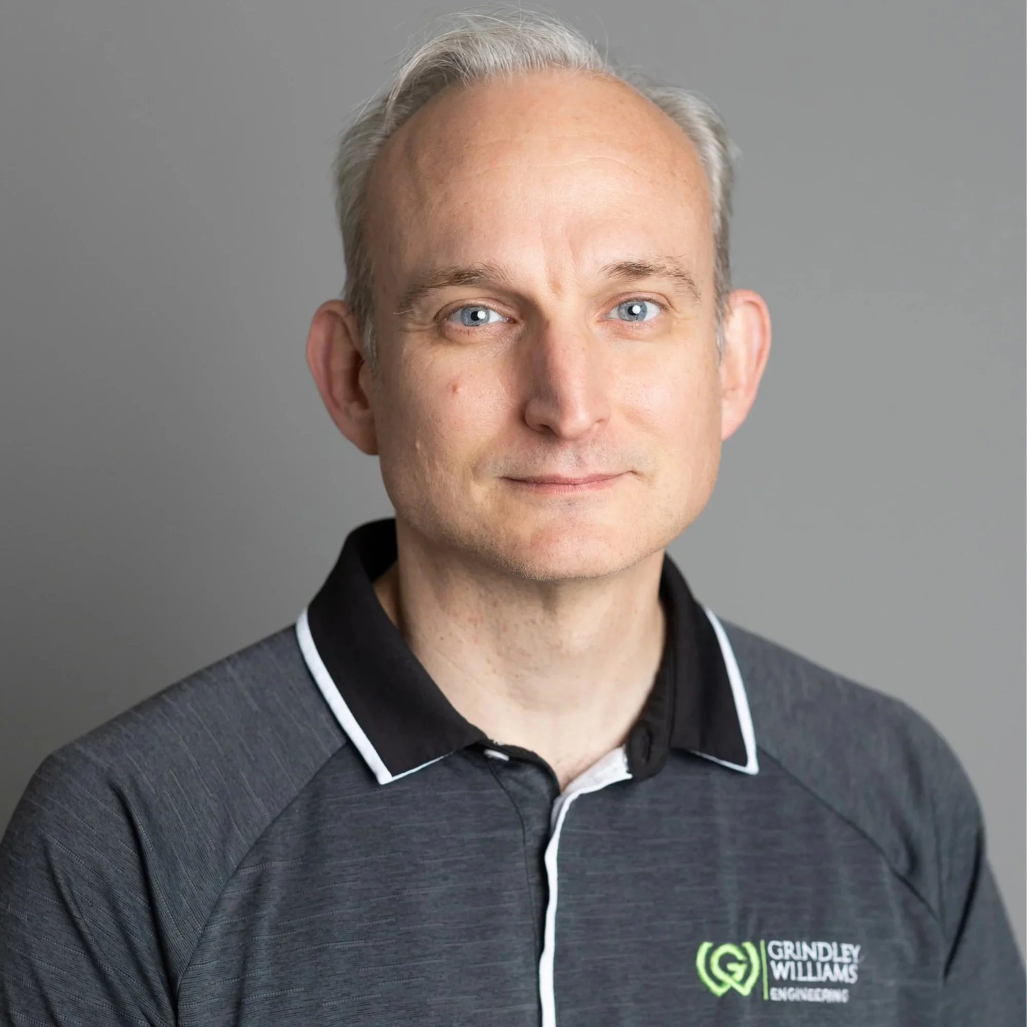 Smiling man in black polo shirt standing against a plain gray background.