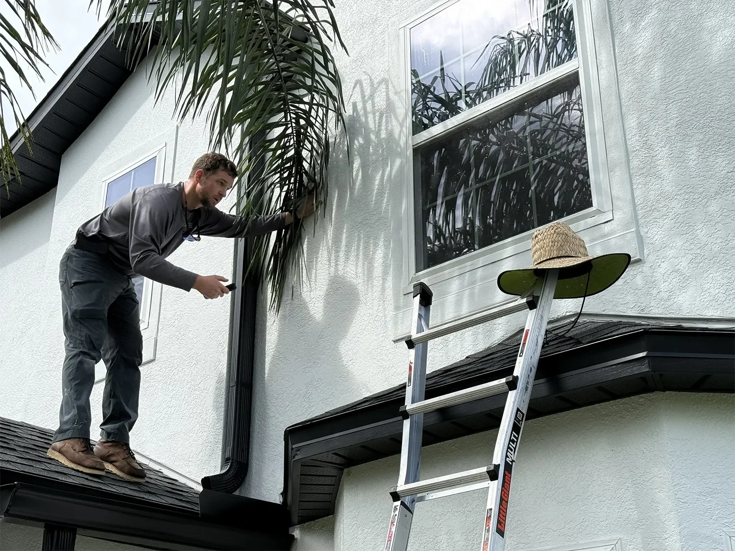 A man standing on a ladder installing or repairing a gutter on a white house. The man is wearing brown shoes, gray pants, and a dark gray shirt. There are trees reflected in a large window next to him and a straw hat resting on the ladder.
