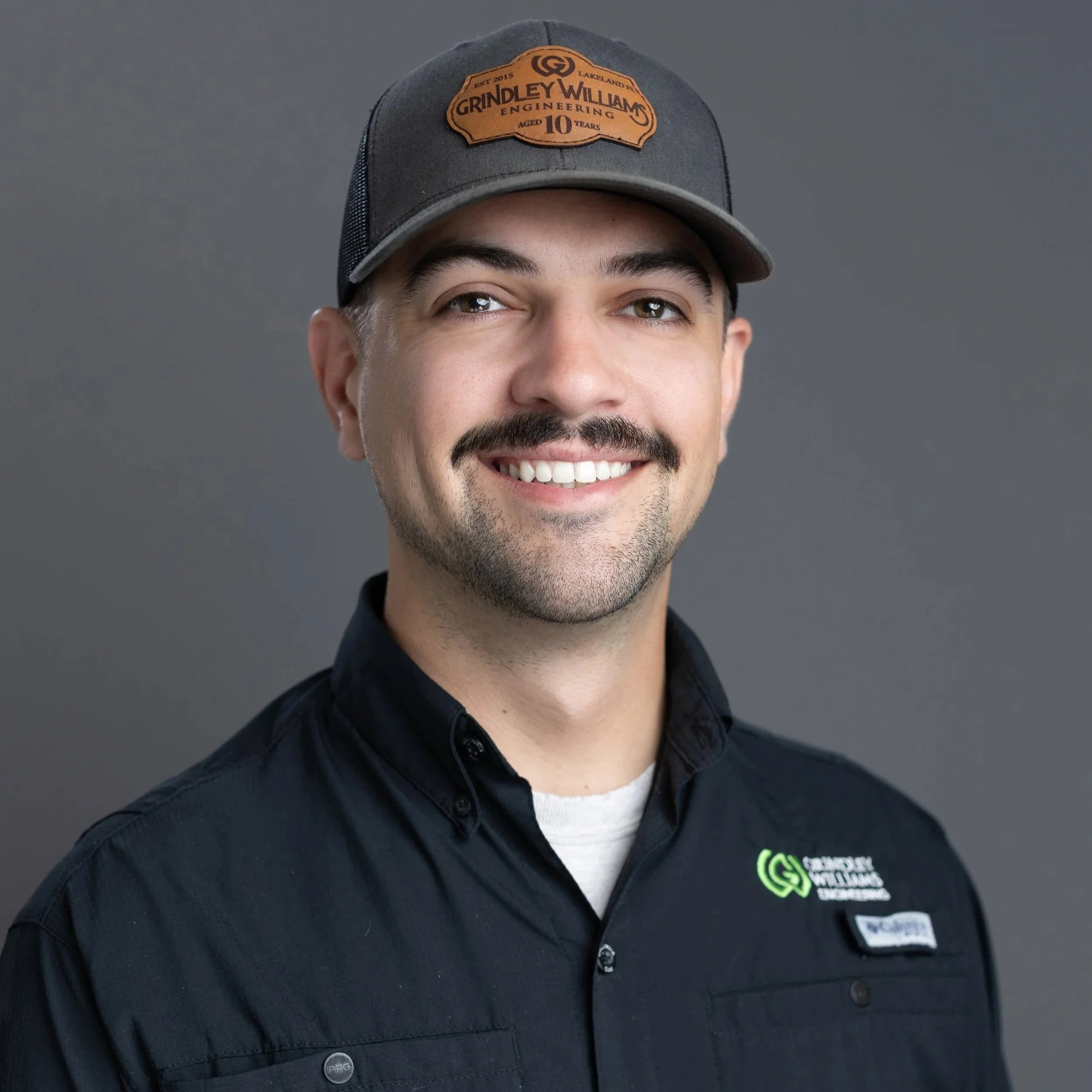 Close-up portrait of a smiling man with dark hair, facial hair, and light skin, wearing a navy blue shirt with a name tag that says "HABIT" on a gray background.