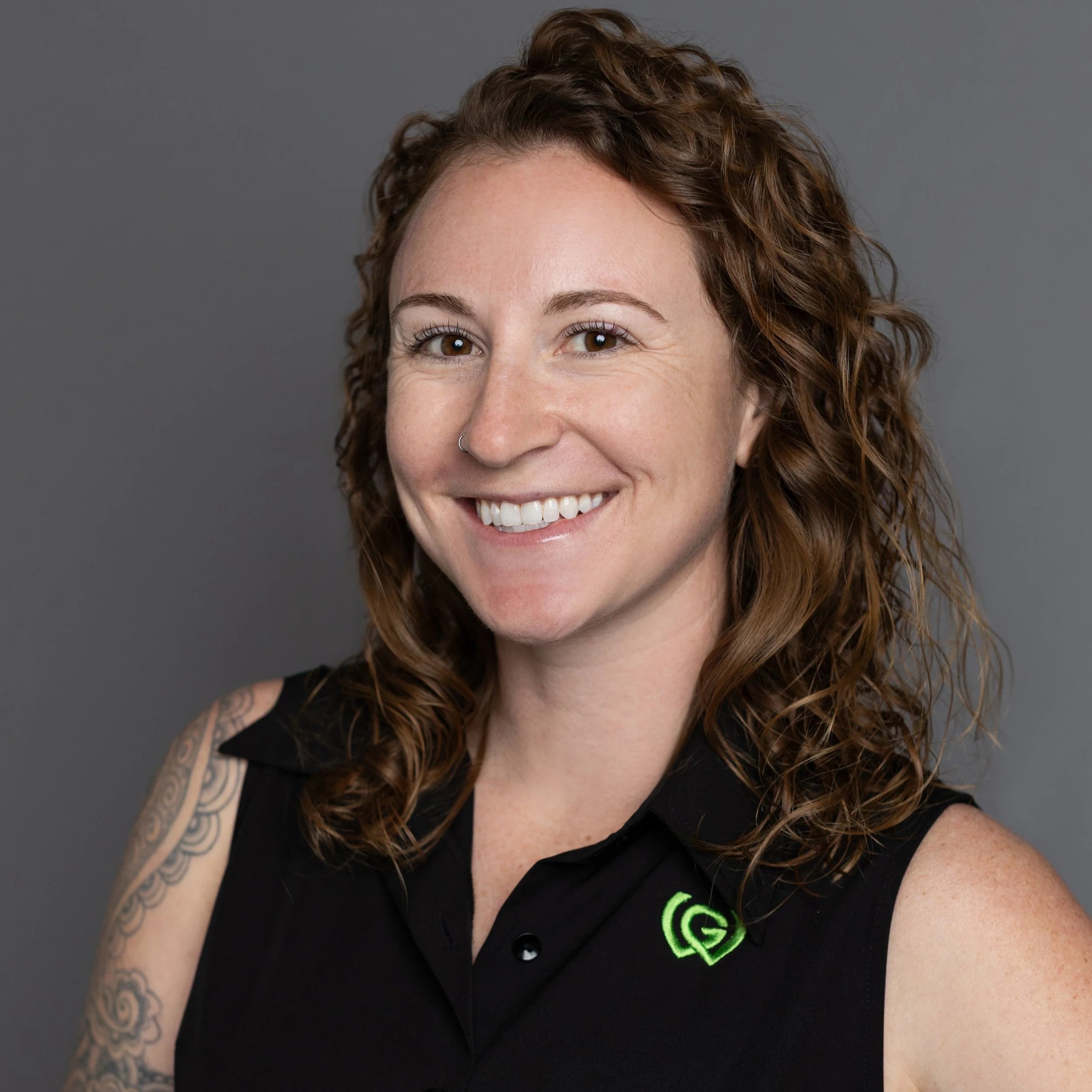 A smiling woman with curly hair, wearing a black sleeveless shirt with a green logo, and showing tattoos on her left arm, standing against a gray background.