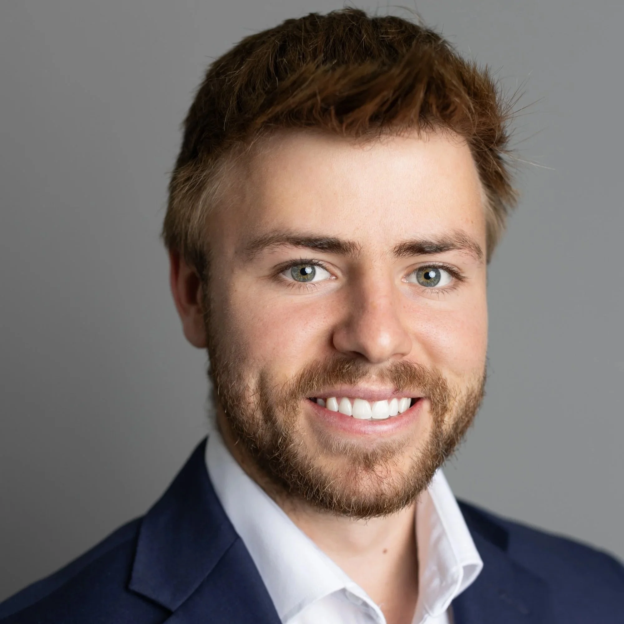 Close-up of a young man with blue eyes, short brown hair, light skin, and a beard, wearing a dark blue suit jacket and a white shirt.
