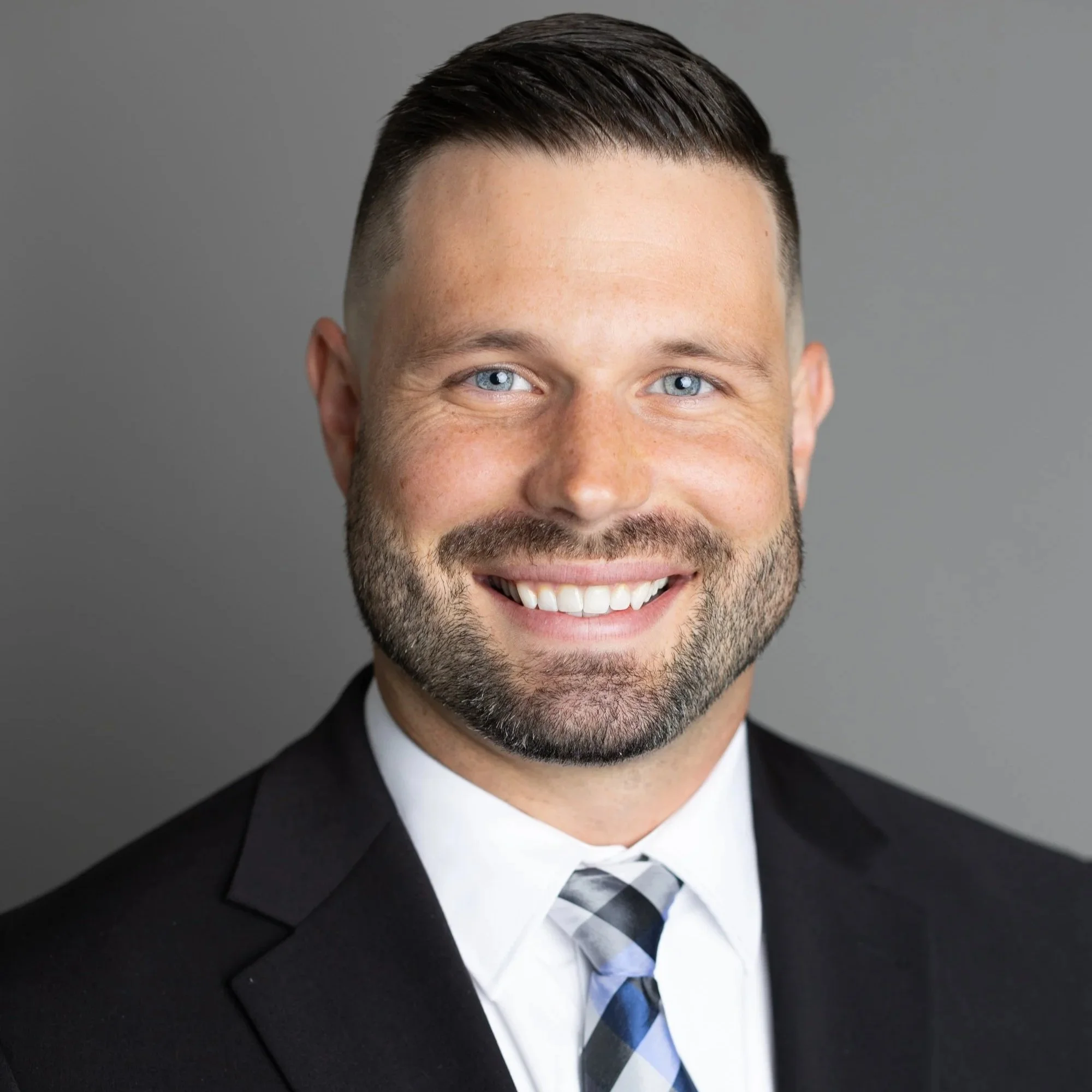 Headshot of a smiling man with blue eyes, dark hair, and a neatly trimmed beard, wearing a black suit, white shirt, and a plaid tie, against a plain gray background.