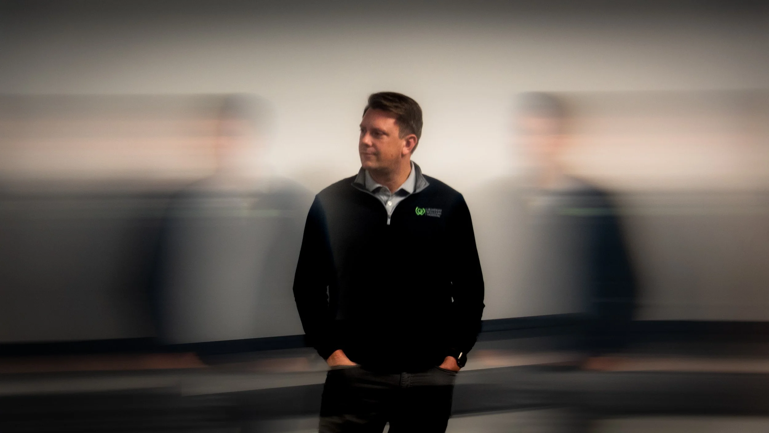 A man standing in front of a blurred background with mirrored images of himself on either side, wearing a black jacket with a logo.