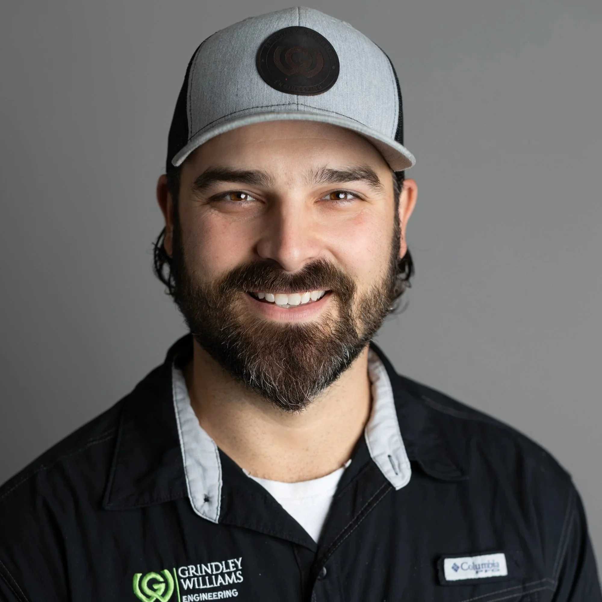 A smiling man with a beard wearing a gray and black cap and a black jacket with embroidery and logos, against a gray background.
