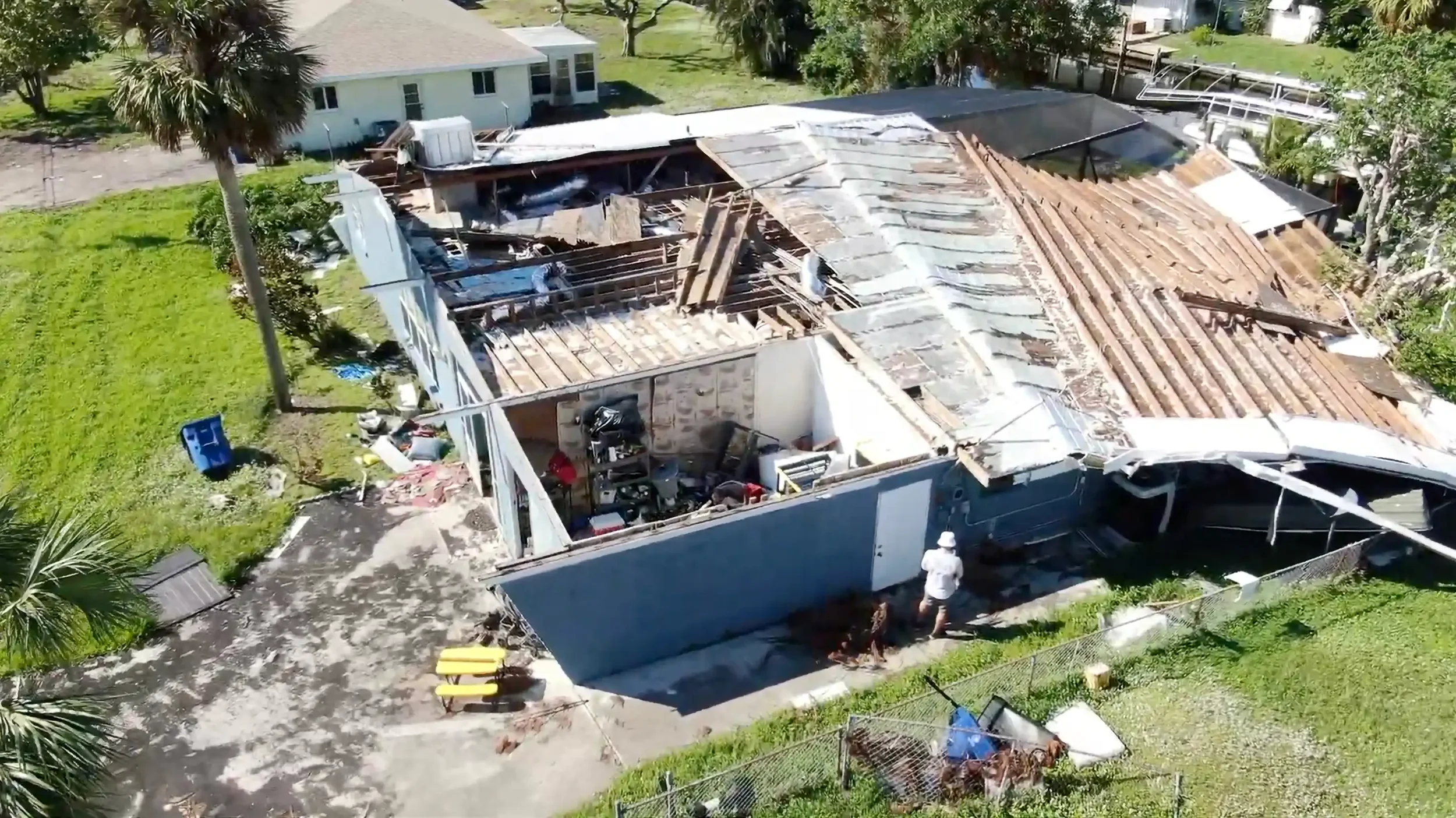 A house with a damaged roof partially collapsed, with people inspecting the damage outdoors. Debris scattered around the yard, surrounded by green grass and trees.