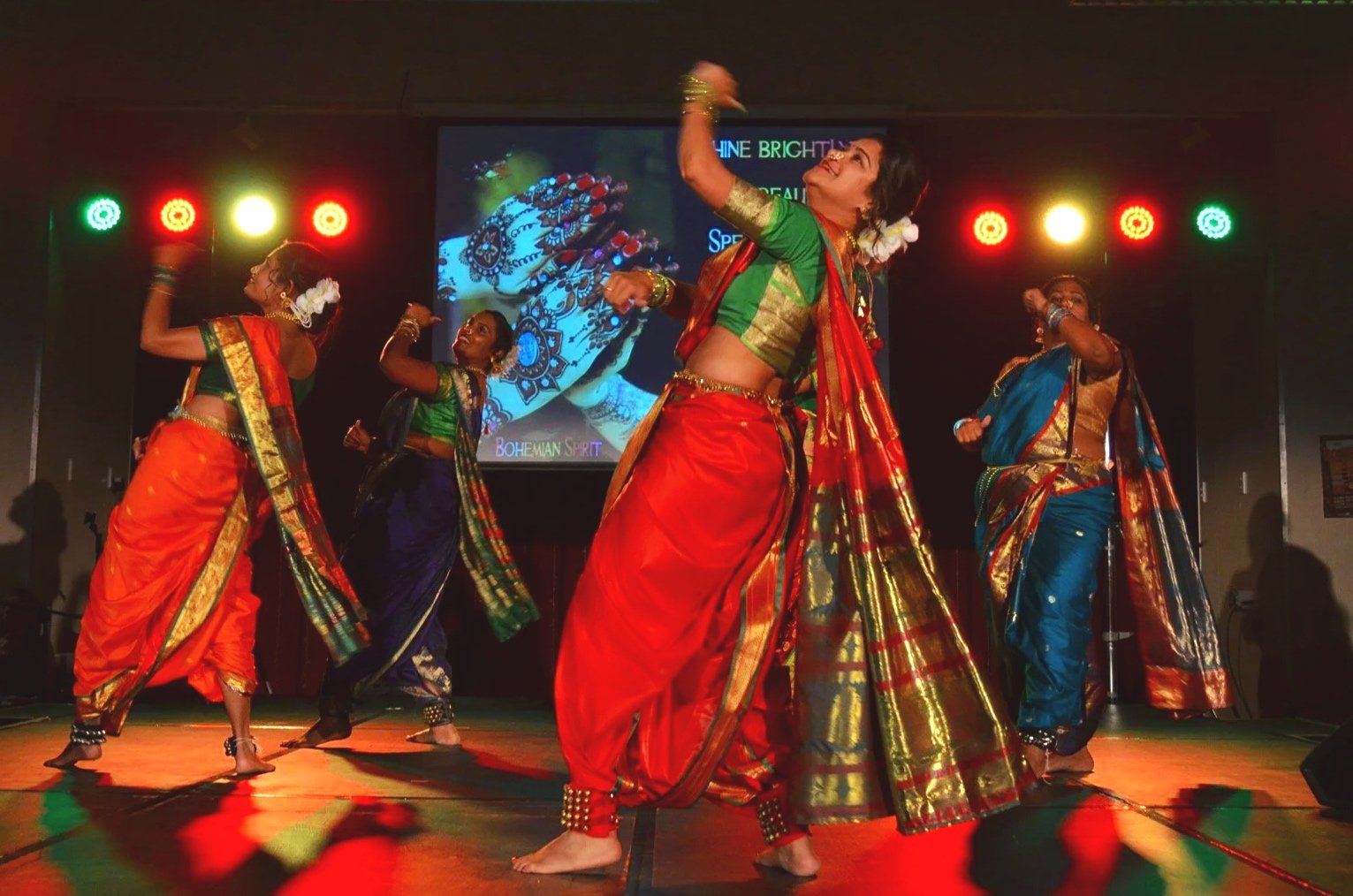 Bollywood Divas in colorful traditional Indian sarees performing a dance on stage with red, green, and yellow stage lights.