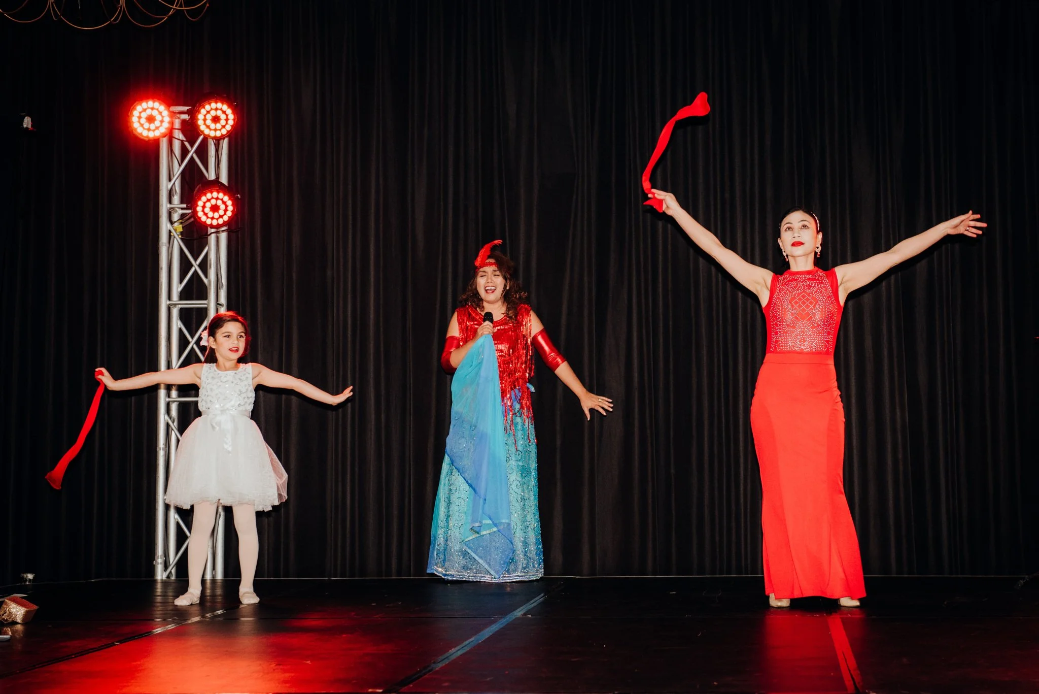 Three performers on stage, representing three generations. A young girl in a white dress with a red ribbon and white tights, a woman in a colorful dress holding a microphone, and another woman in a red dress with her arms raised, holding a red scarf.