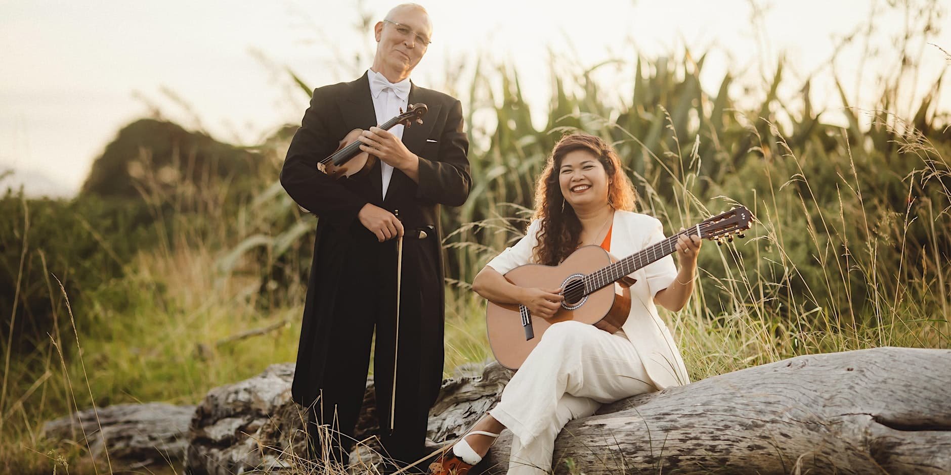 Crescendo members Albert and Lyly holding their violin and guitar, sitting on a fallen log in a grassy outdoor setting in New Plymouth, New Zealand.