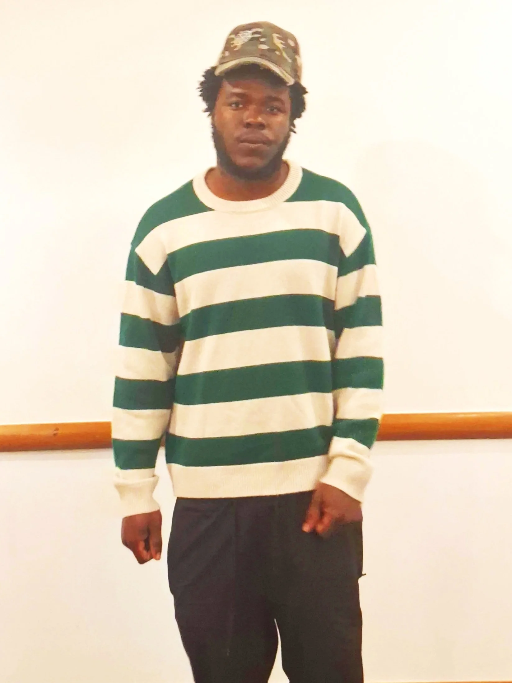 Singer Sonylekxy, wearing a camouflage cap and a striped green and white sweater, standing against a plain light-colored wall.