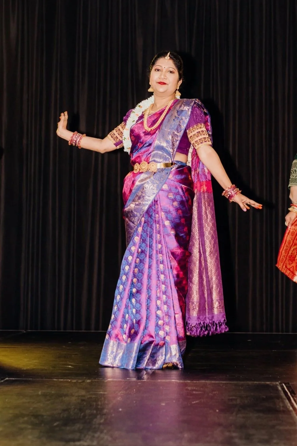 A dazzlingly elegant Indian model wearing a vibrant purple and pink sari adorned with gold jewelry and floral garlands.