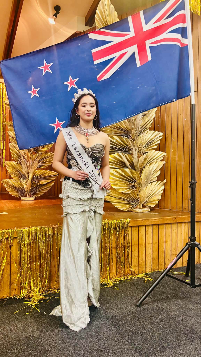 Kate Lucio wears her Miss Taranaki 2026 sash, crowned and elegant in front of the New Zealand flad
