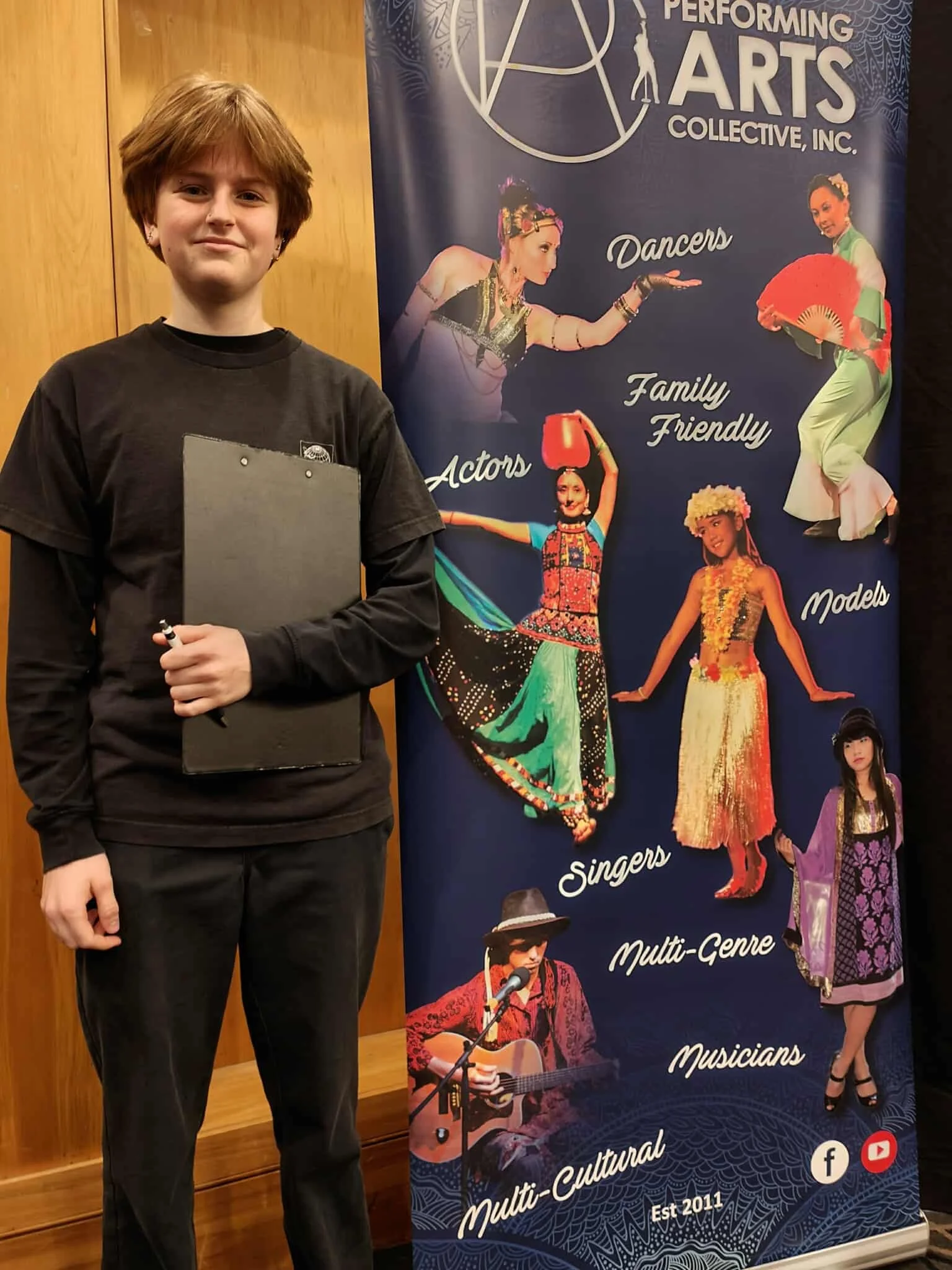 A PAC volunteer standing next to a banner with the words 'Performing Arts Collective, Inc.' and images of dancers, actors, singers, multi-cultural performers, and musicians.