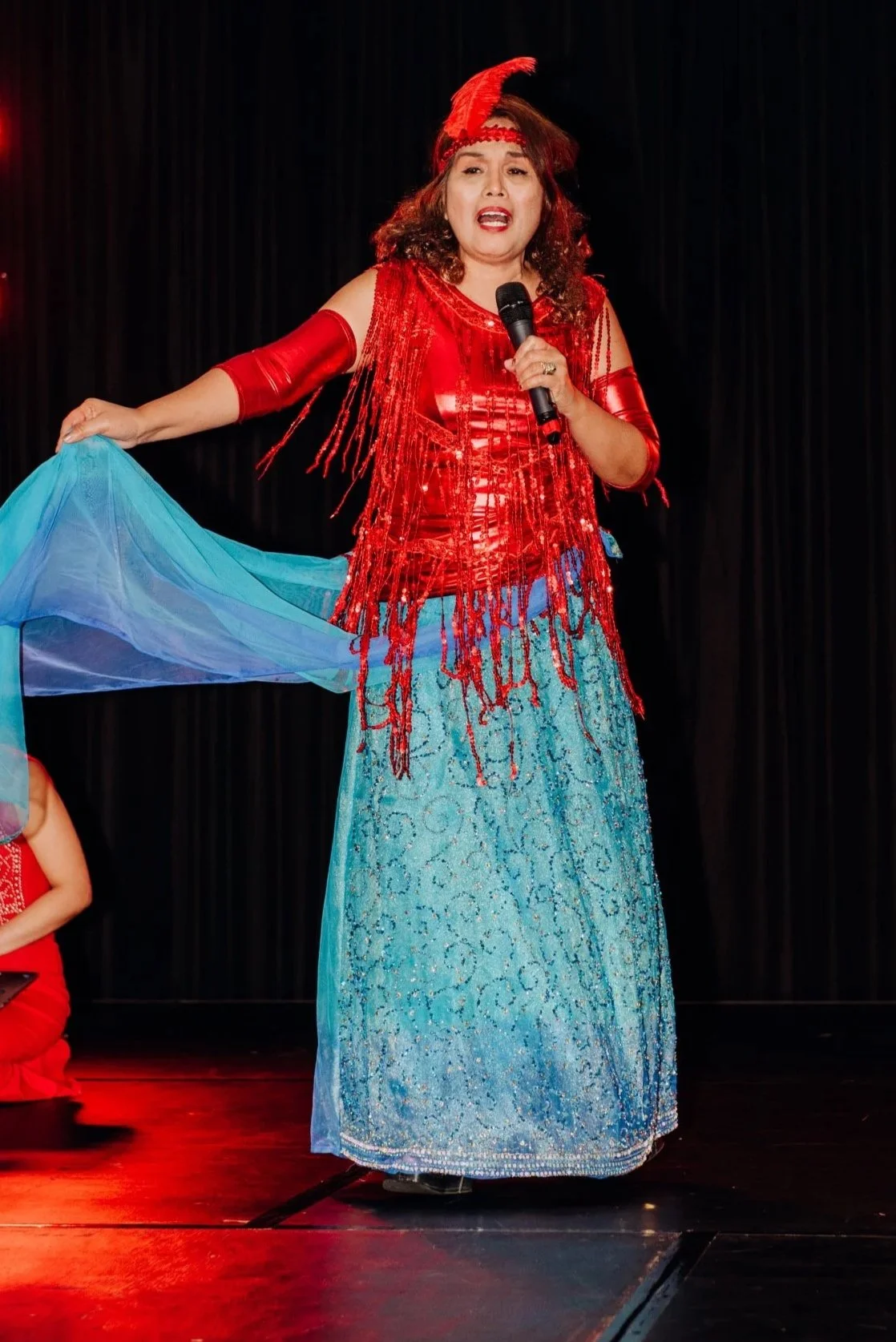 Emily Julian dressed in colorful, elaborate costume performing on stage with a black curtain background, holding a microphone, and wearing a red feathered headband and a flowing blue skirt.