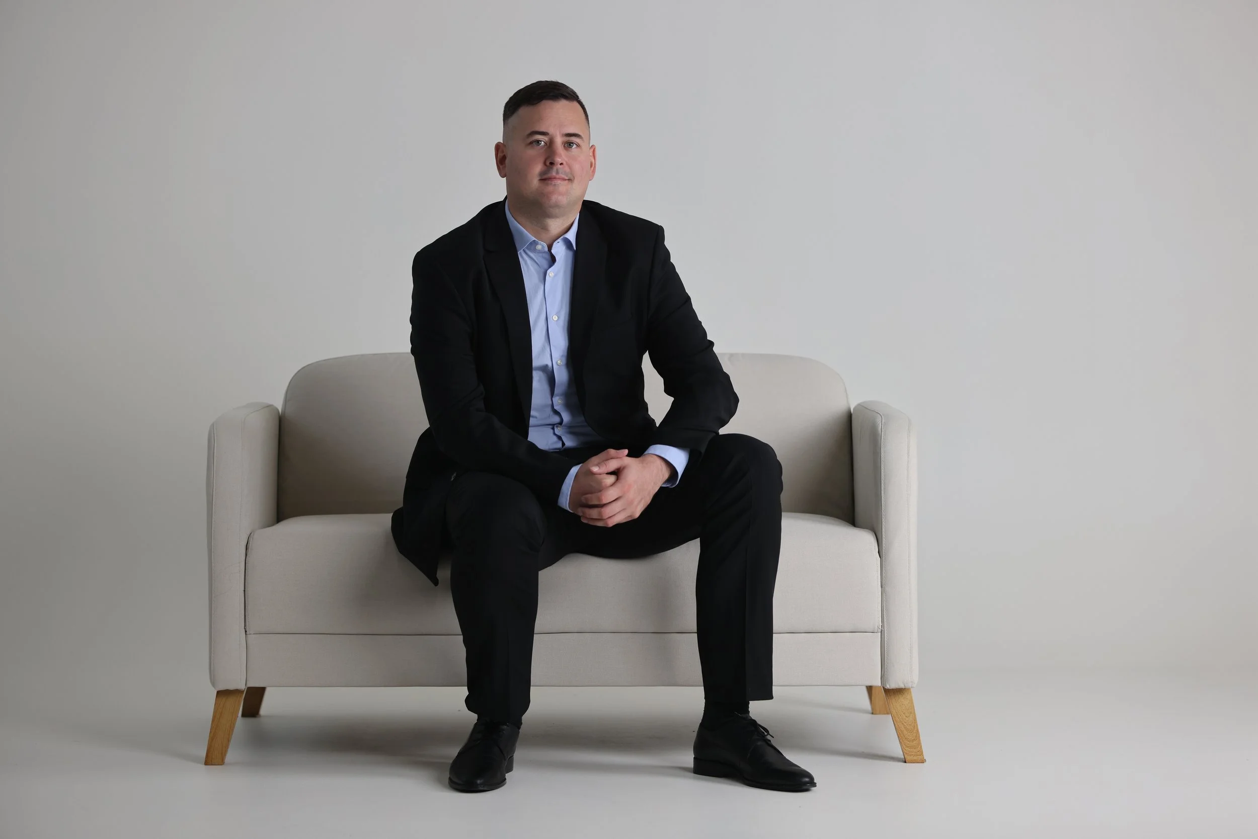 A man in a black suit and blue shirt sitting on a cream-colored sofa against a plain white wall.