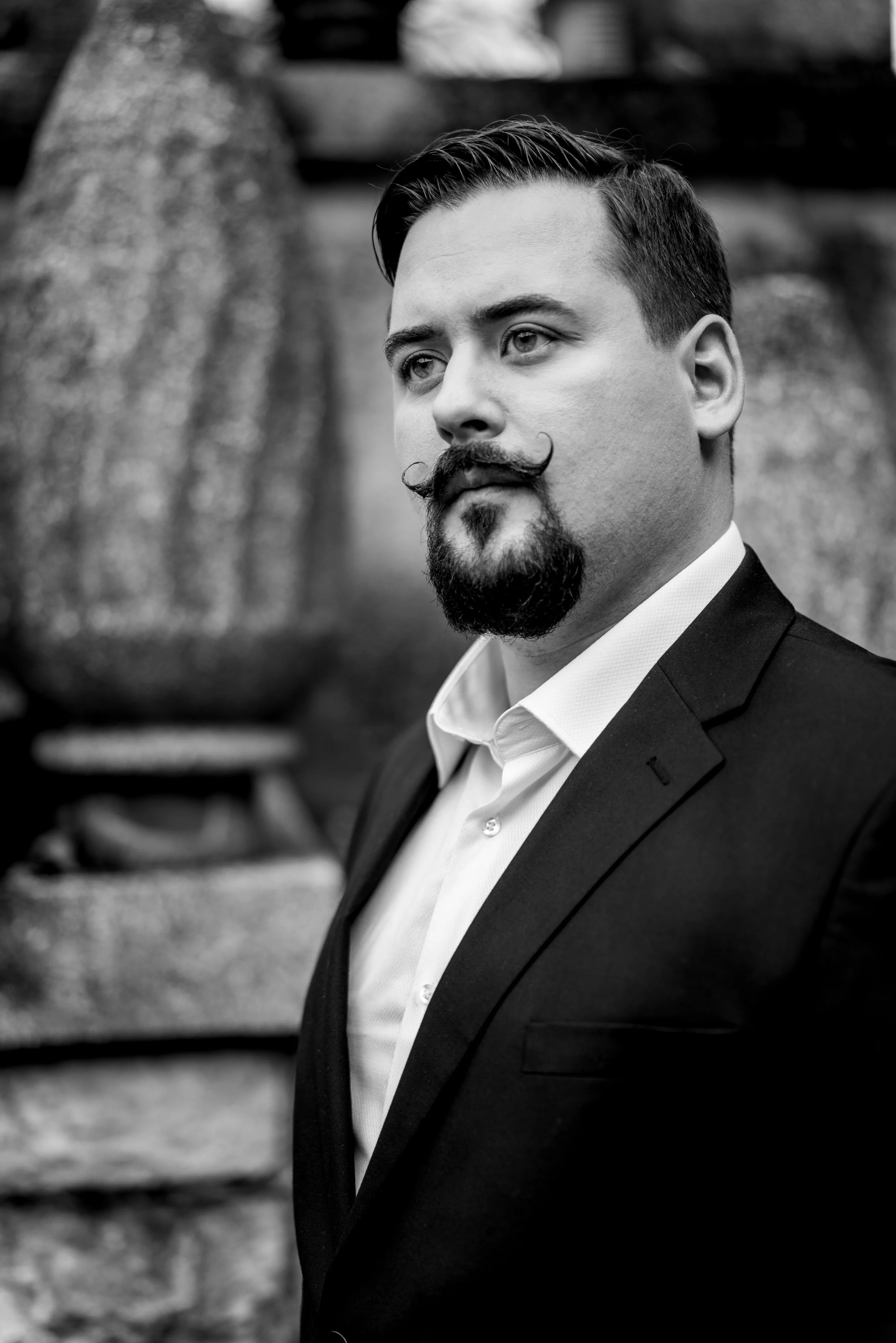 A black-and-white portrait of a man in a suit with a styled mustache and goatee, standing outdoors in front of large rocks.