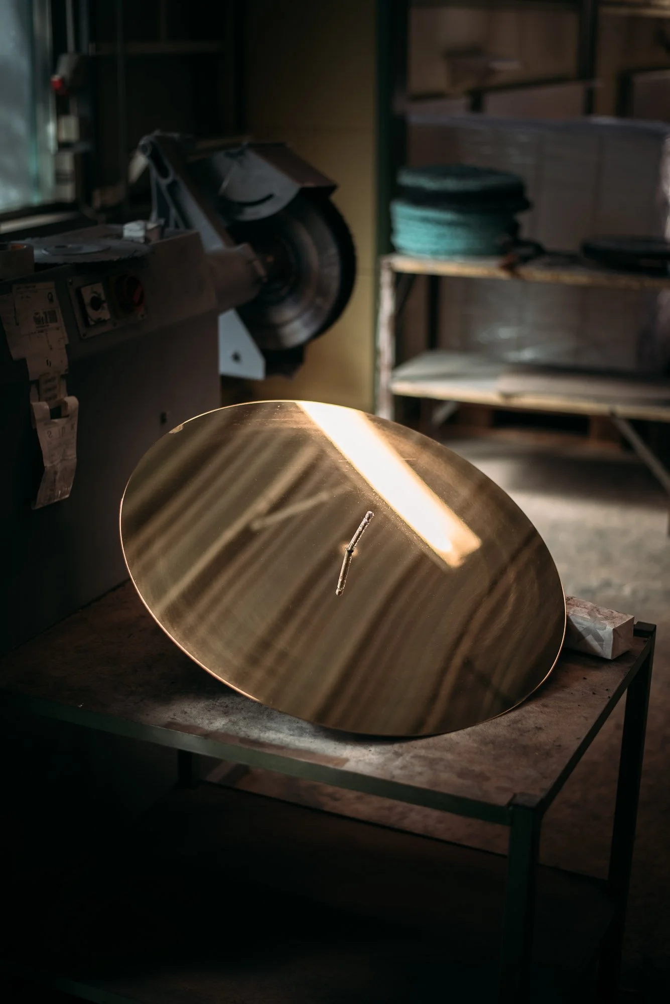 A shiny, reflective, oval-shaped metal disc resting on a work table in a workshop, with various tools and equipment visible in the background.