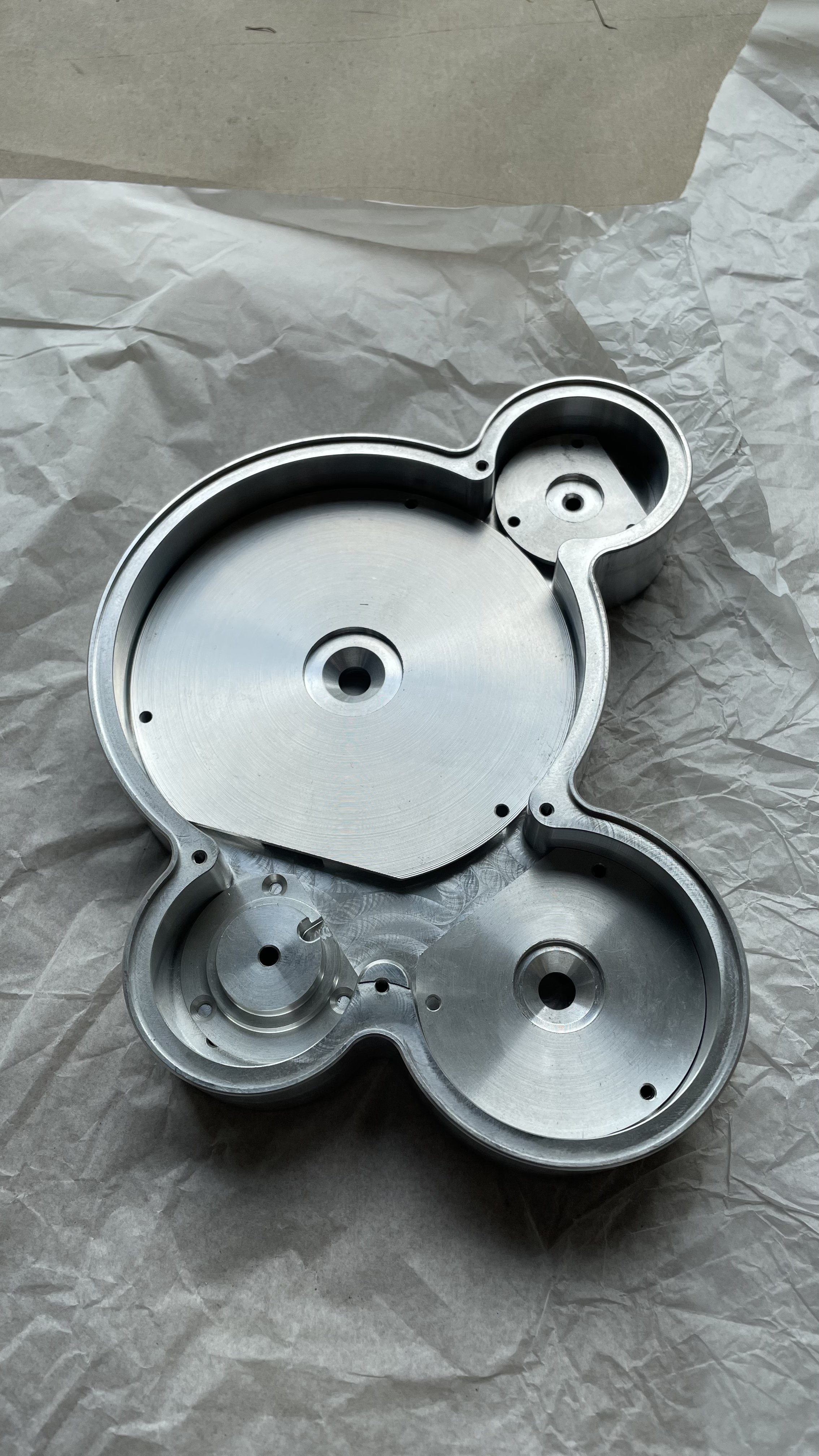 A metallic, multi-compartment, engraved aluminum part with multiple circular sections and small holes, resting on crumpled tissue paper.