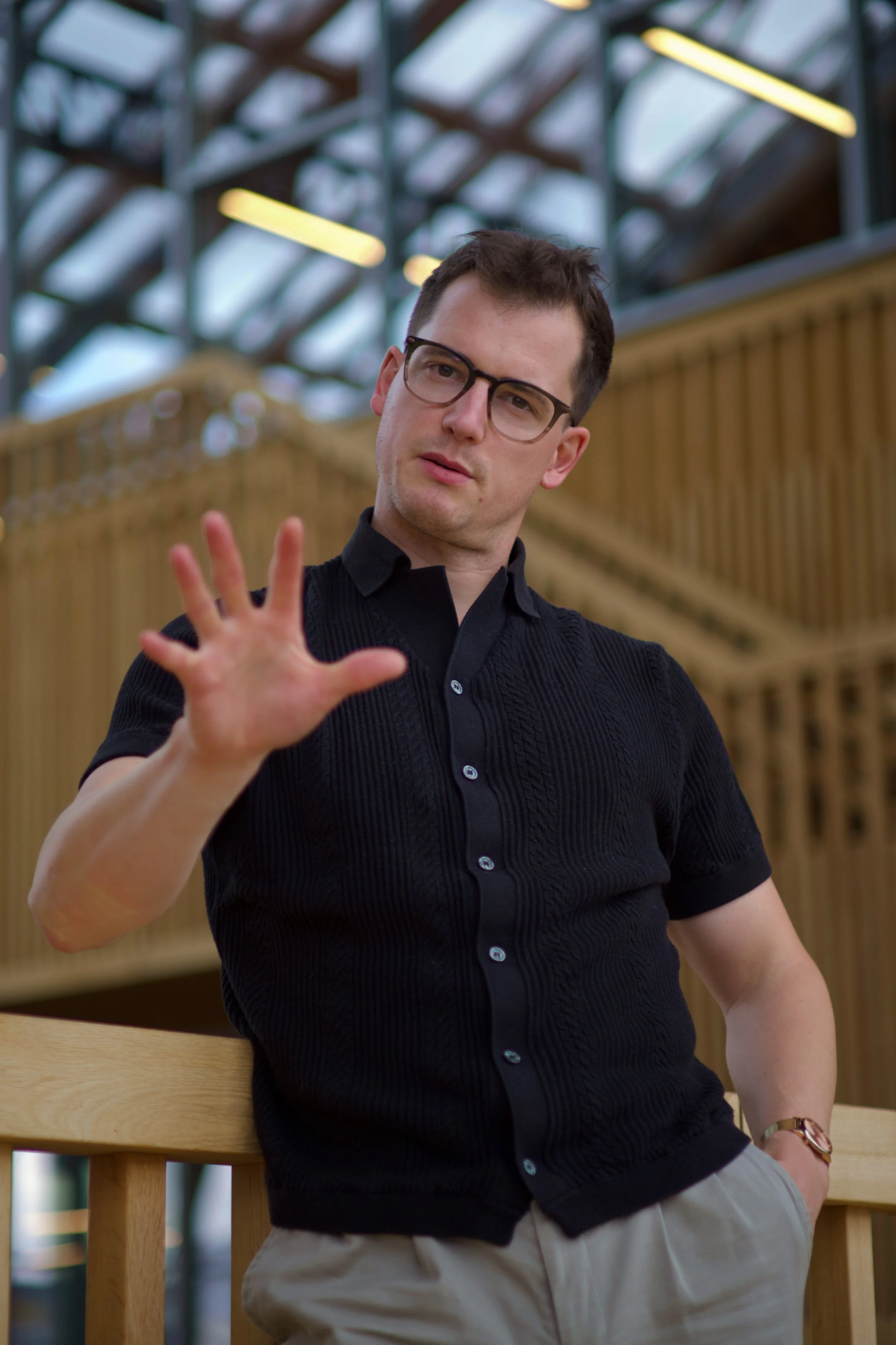 A man with short dark hair, glasses, wearing a black short-sleeved shirt and beige pants, standing outdoors near a wooden railing with a blurred background of metal framework and yellow lights.