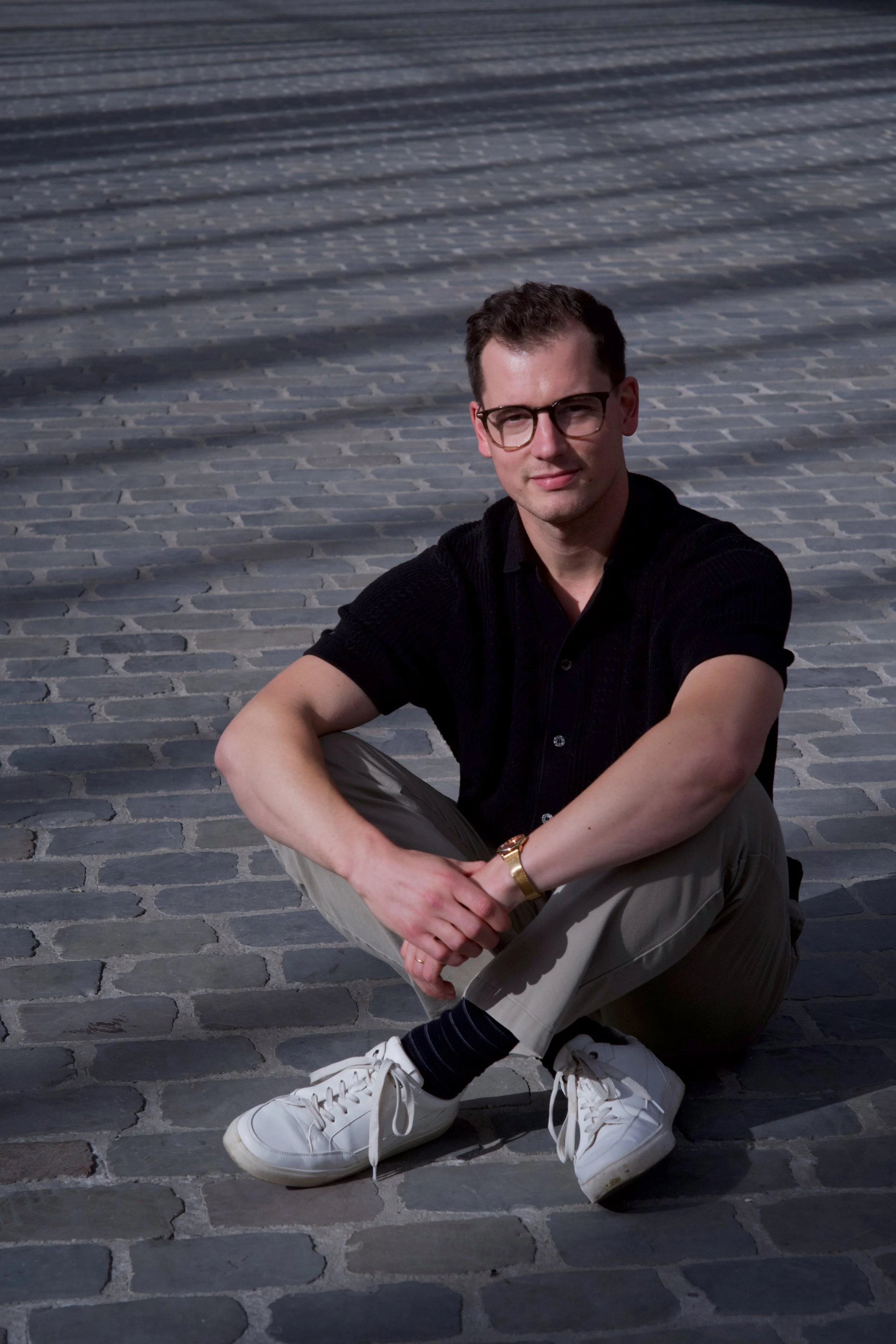 A young man with short dark hair and glasses sitting on a cobblestone street, wearing a black shirt, beige pants, white sneakers, black socks, and a gold watch.