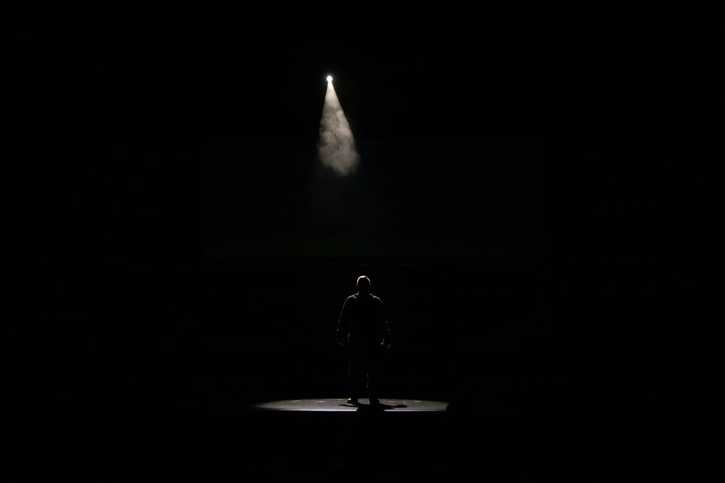 A person standing on a stage in front of a dark background, illuminated by a spotlight from above.