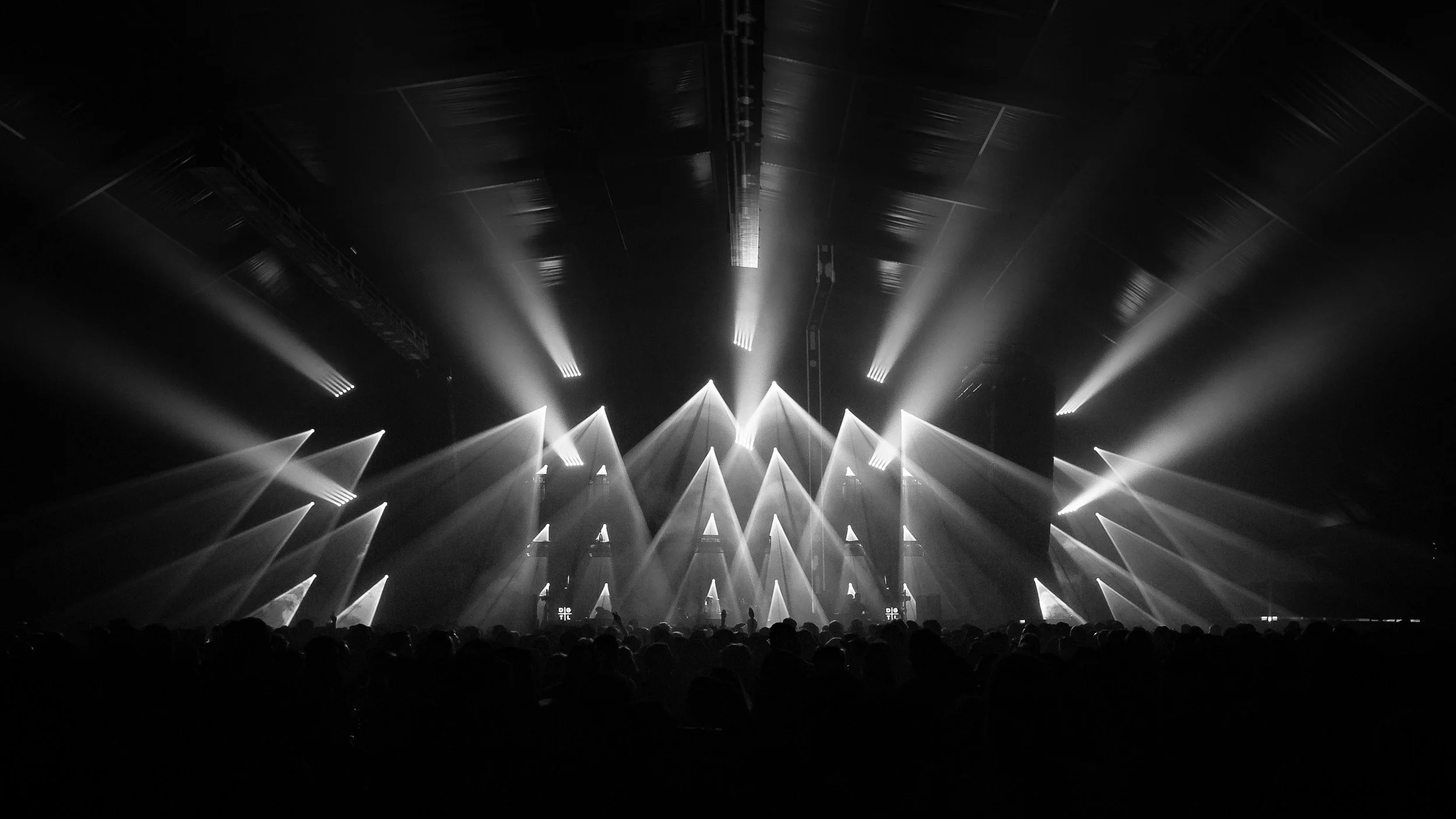 Black and white photograph of a concert with stage lighting, beams, and an audience in silhouette.