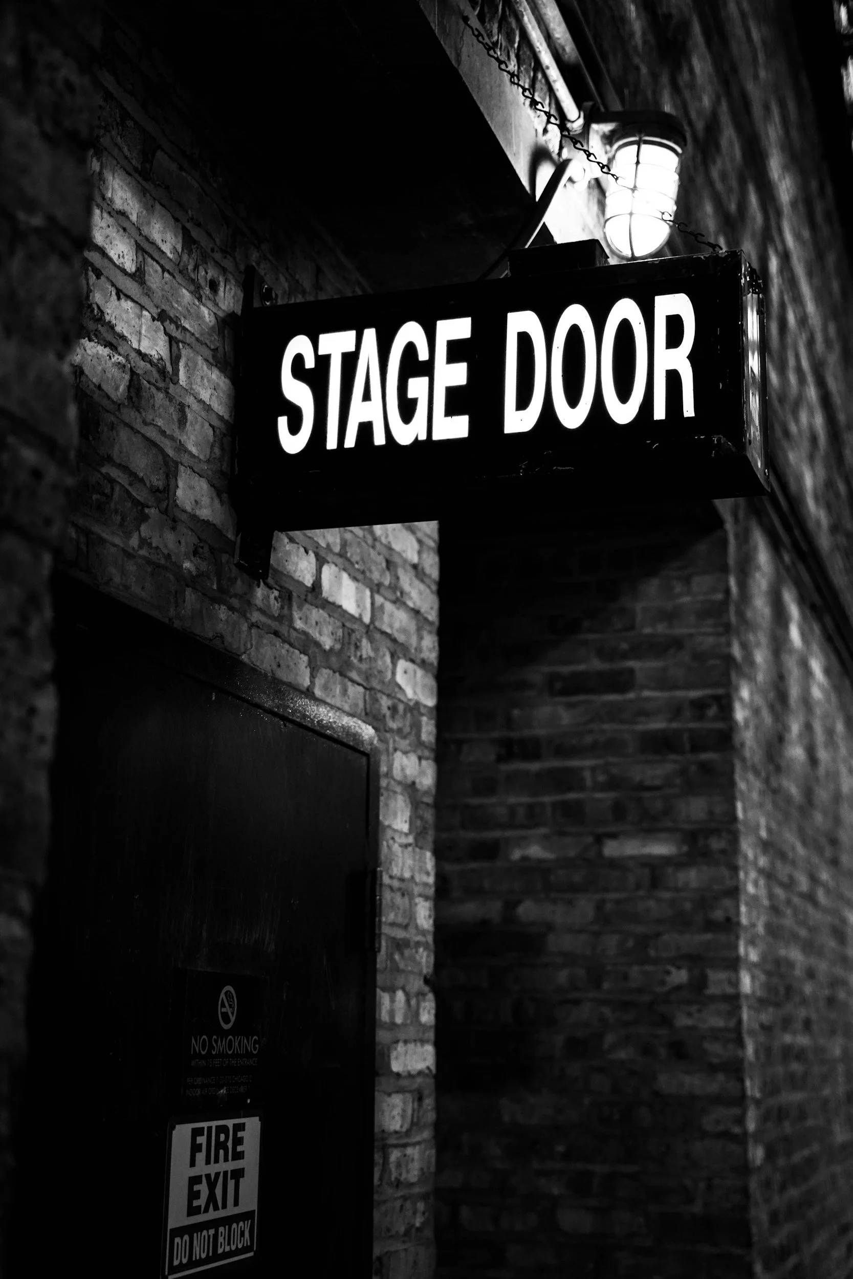 Black and white photograph of a lit sign that says 'STAGE DOOR' attached to a brick wall, with a light fixture above it.