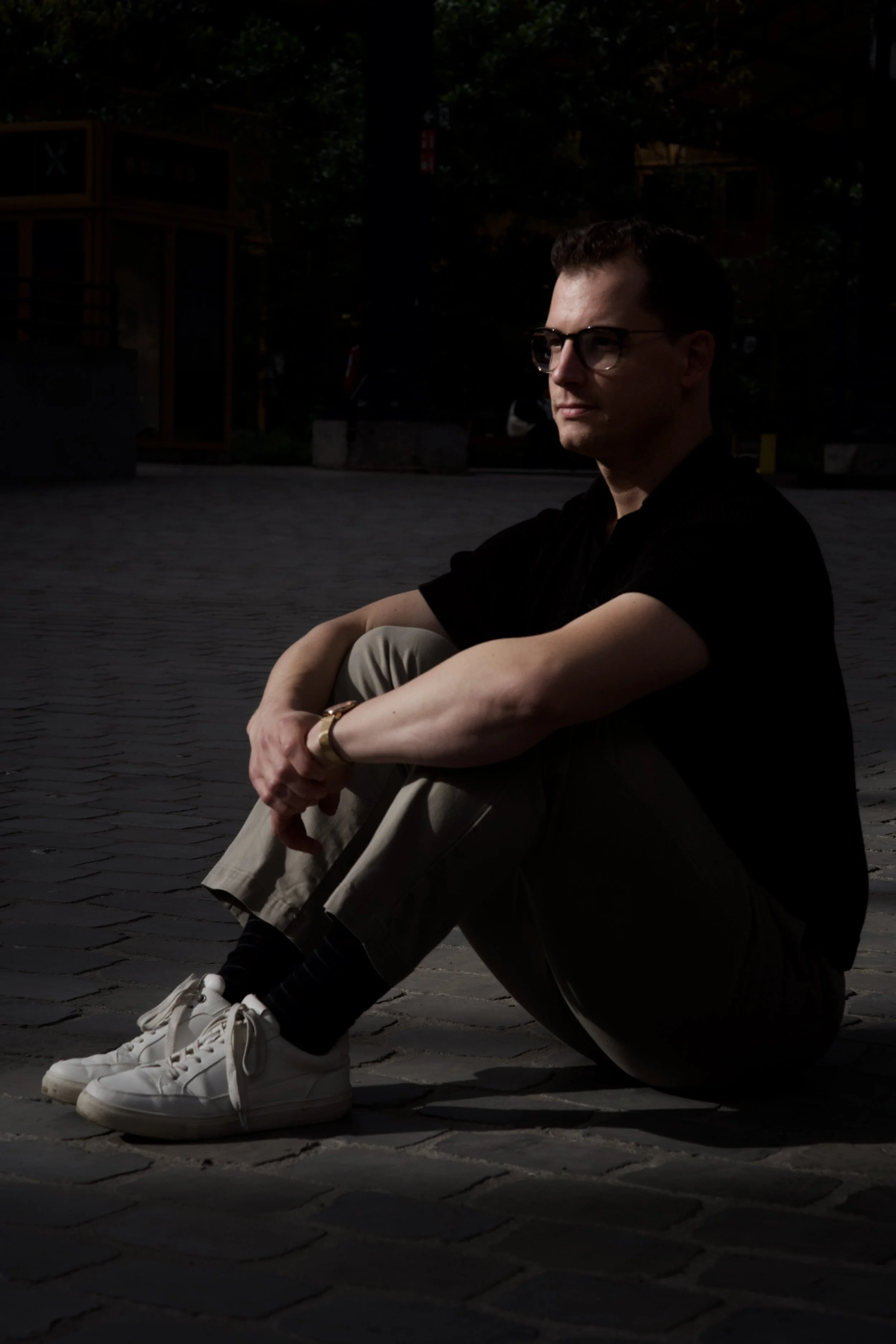A man with glasses, dark hair, sitting outdoors on pavement, wearing a black shirt, beige pants, white sneakers, and a gold watch, with a background of trees and buildings, shot in low light.