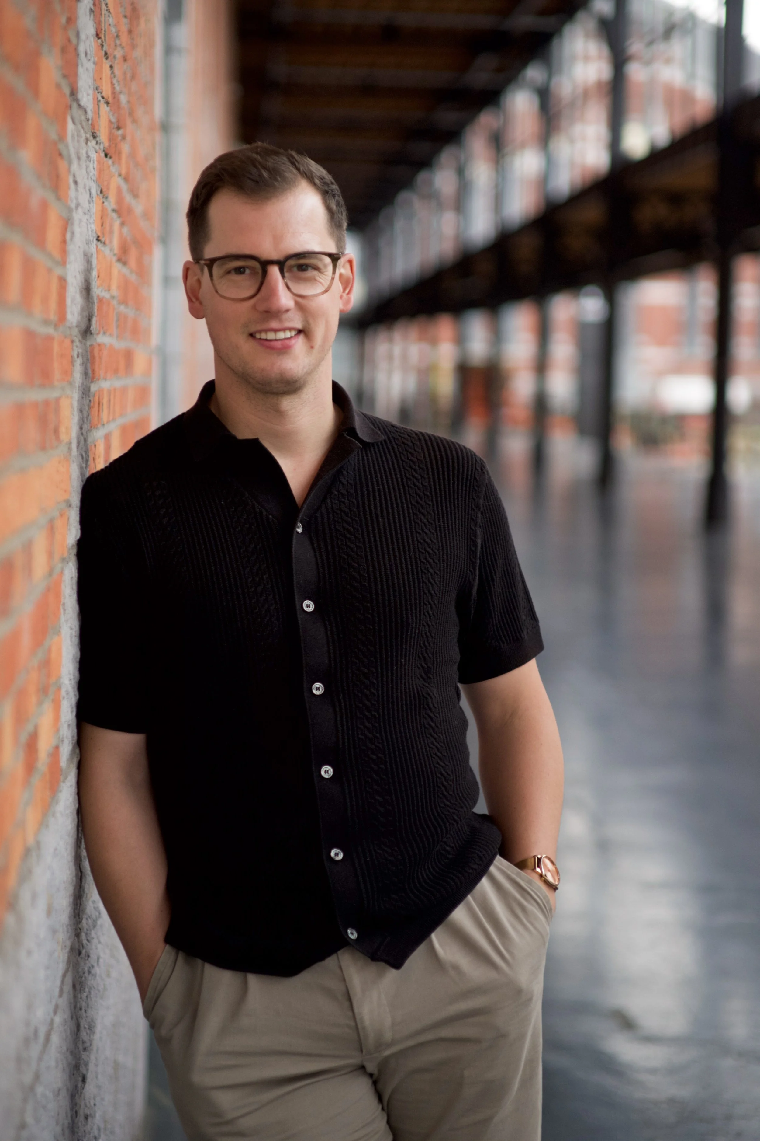 A young man with short dark hair, wearing glasses, a black short-sleeve button-up shirt, and beige pants, leaning against a brick wall in an outdoor alleyway during daytime.