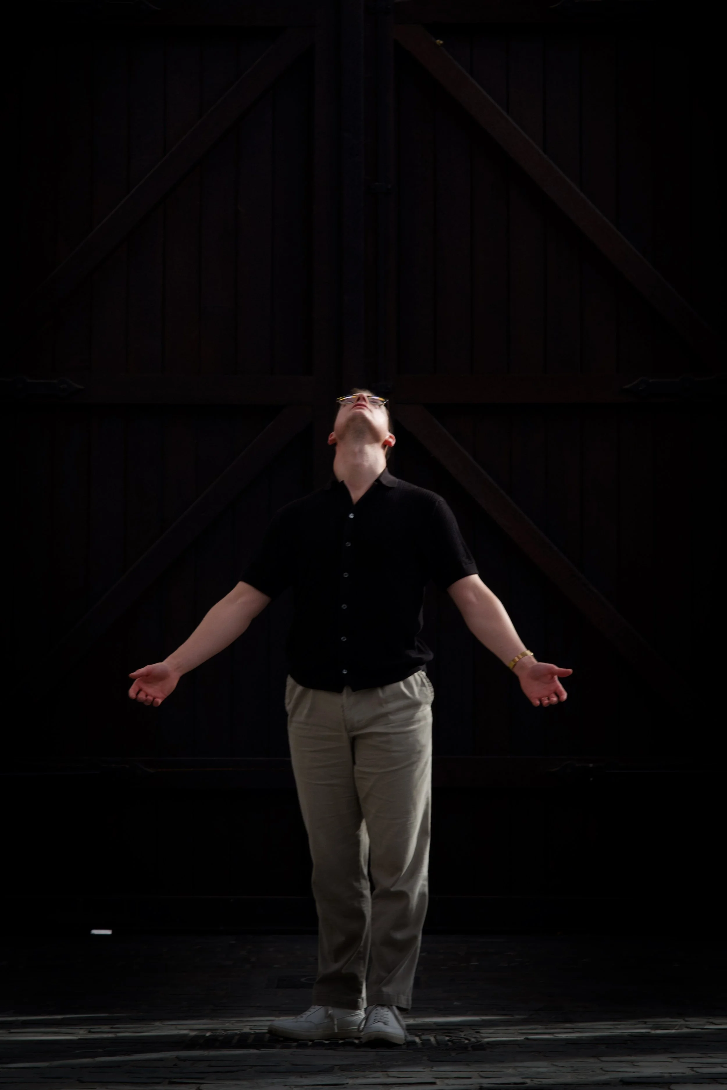 A man wearing glasses, a black shirt, and beige pants stands with arms outstretched in front of a dark wooden barn door, looking upward.