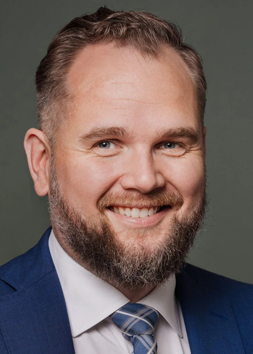 Portrett av en mann med lyst hår, skjegg og blå øyne, iført en blå dress, hvit skjorte og blå-stripet slips, smilende.