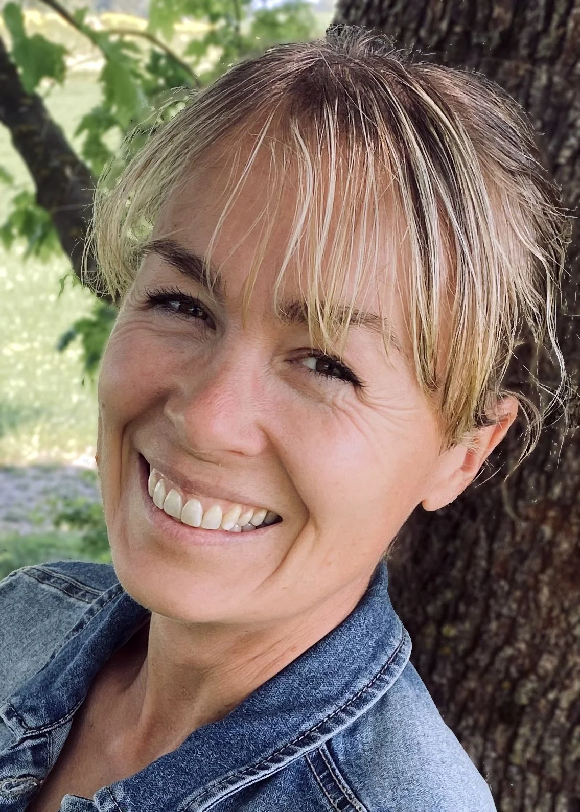Nærbilde av en smilende kvinne med kort blond frisyre og denimjakke, stående ved et tre utendørs med grønt løvverk i bakgrunnen.