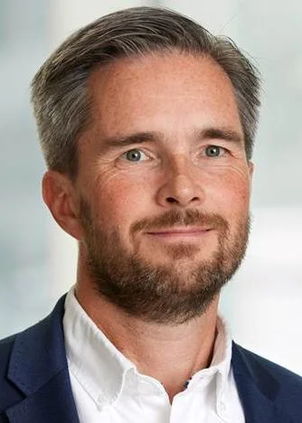 Portrettbilde av en mann med kort, grå hår og skjegg, iført en hvit skjorte og mørkeblå blazer, poserer innendørs.