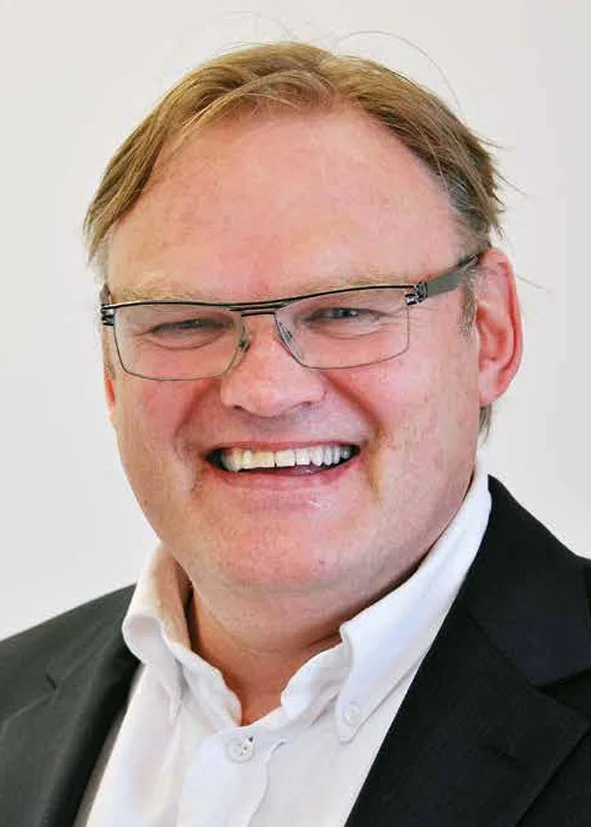 En smilende mann med lys hud, blond hår, briller og iført sort blazer og hvit skjorte.