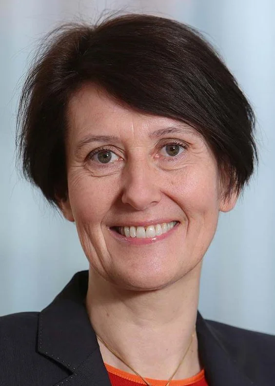 Nærbilde av en smilende kvinne med kort mørk hår, iført mørk blazer og rød topp, mot en lys bakgrunn.