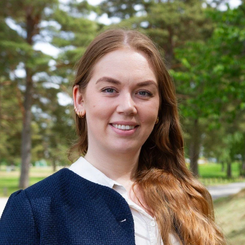 En smilende ung kvinne med langt, bølget, brunt hår, ikledd en hvit skjorte og mørk blazer, står utendørs i en park med grønne trær i bakgrunnen.