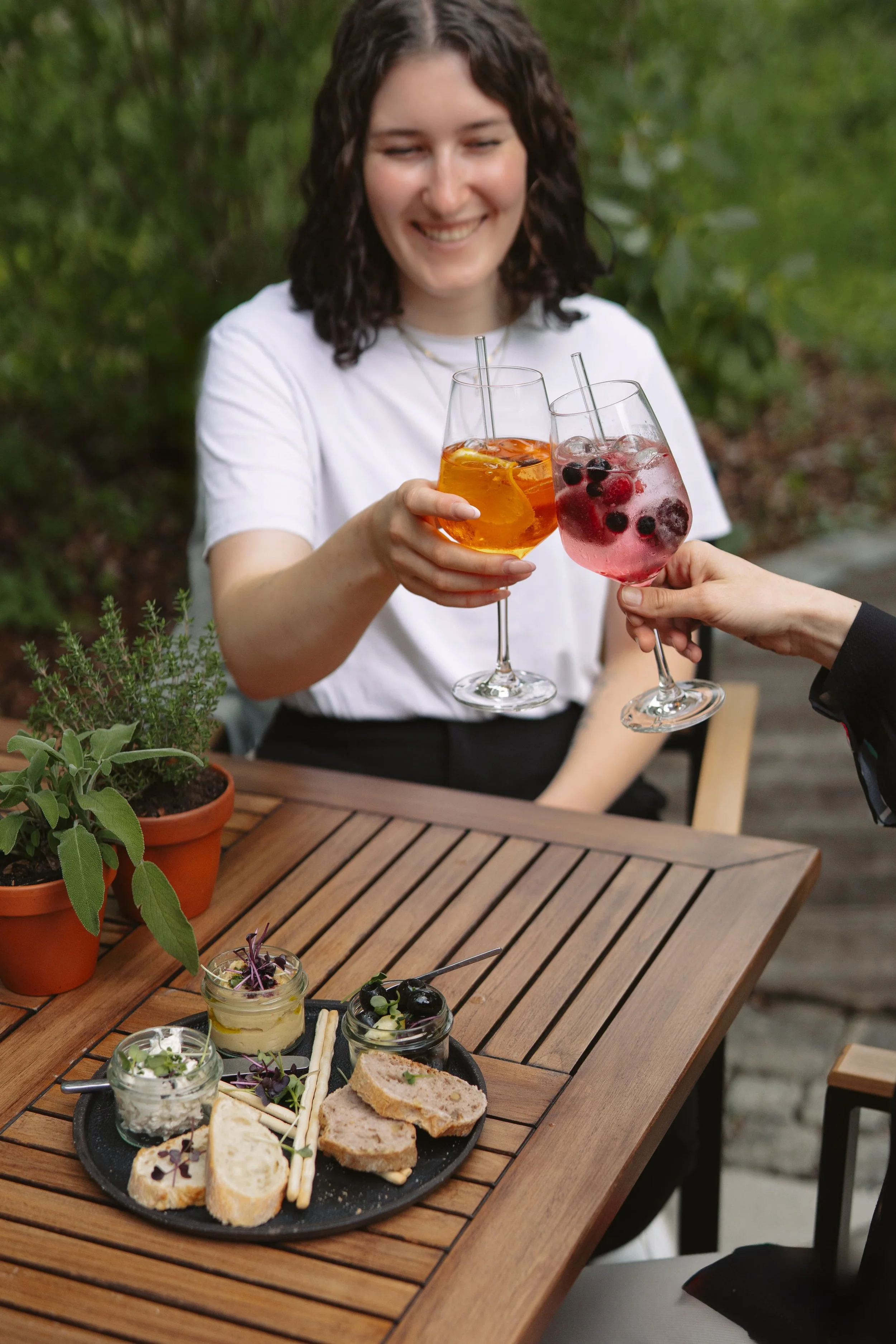 Zwei Frauen stoßen mit bunten Cocktails in einem Garten an, auf dem Tisch sind kleine Vorspeisen, Kräuter und Pflanzen