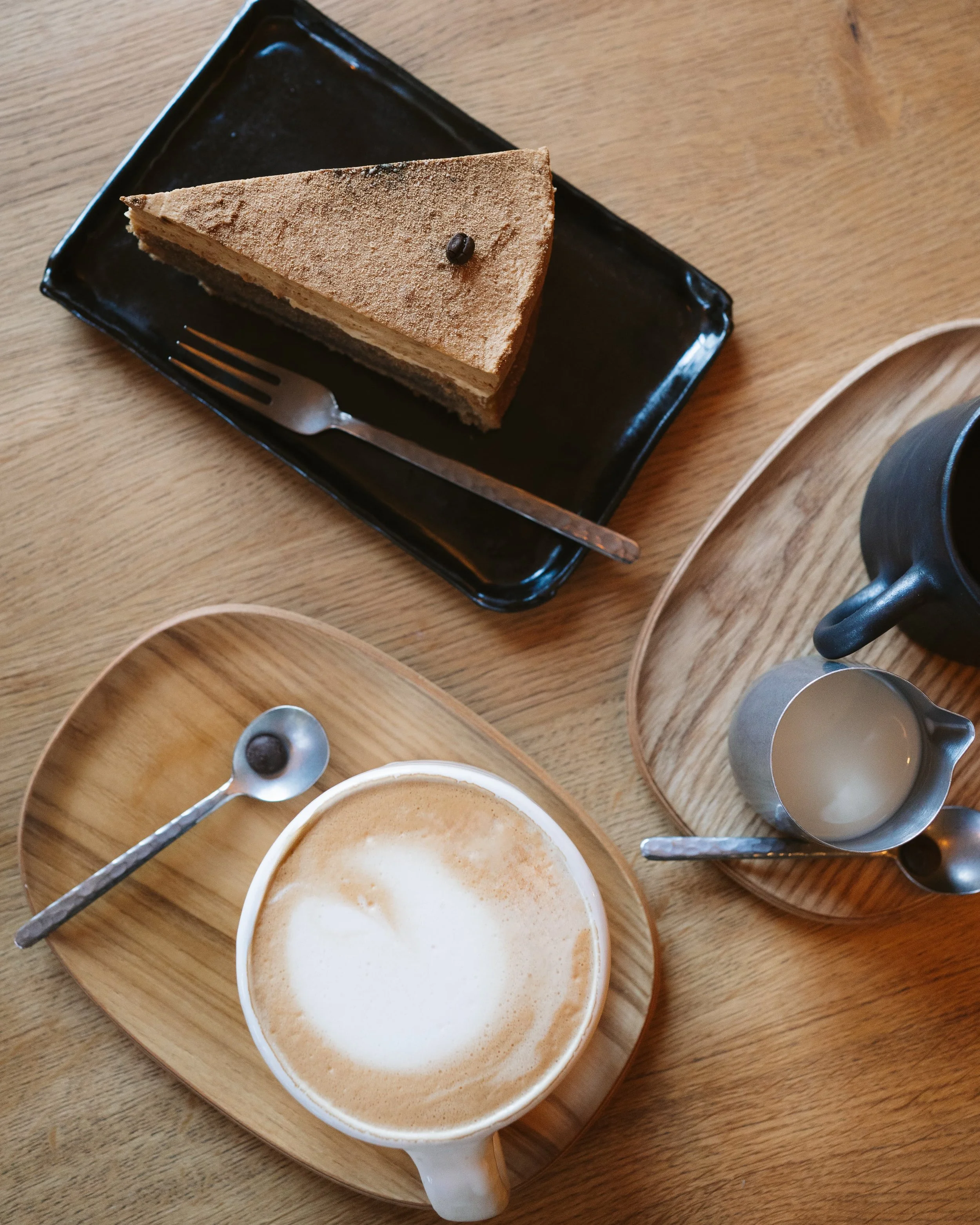 Ein Stück Tiramisu-Kuchen auf einem schwarzen Teller, eine Tasse Cappuccino mit Milchschaum auf einem Holztablett, sowie eine kleine Kanne Milch und andere Tassen auf einem Holztablett.
