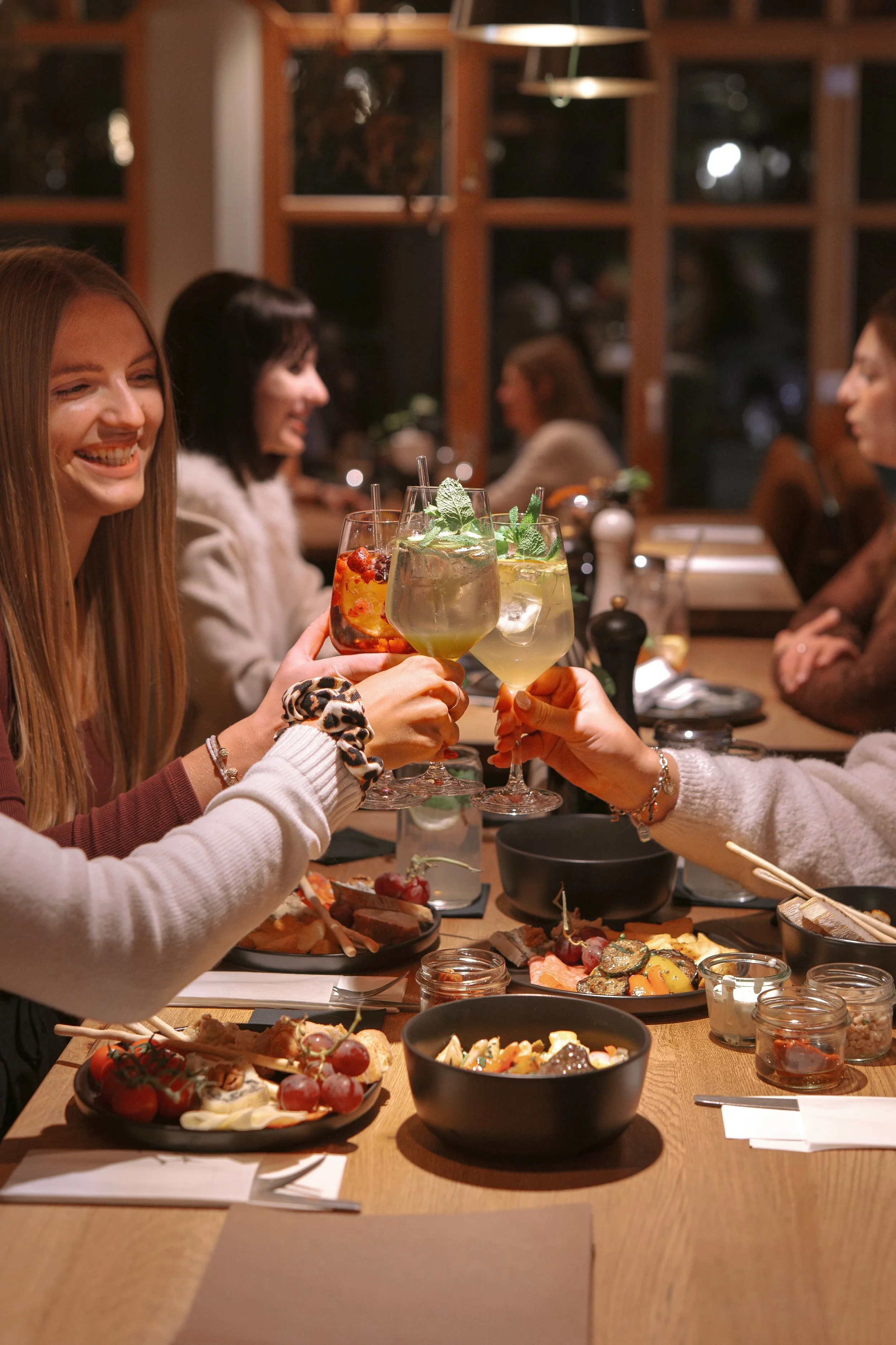 Menschen sitzen an einem Tisch in einem Restaurant, stoßen mit verschiedenen Cocktails an, es gibt verschiedene Speisen wie Früchte und Vorspeisen auf dem Tisch