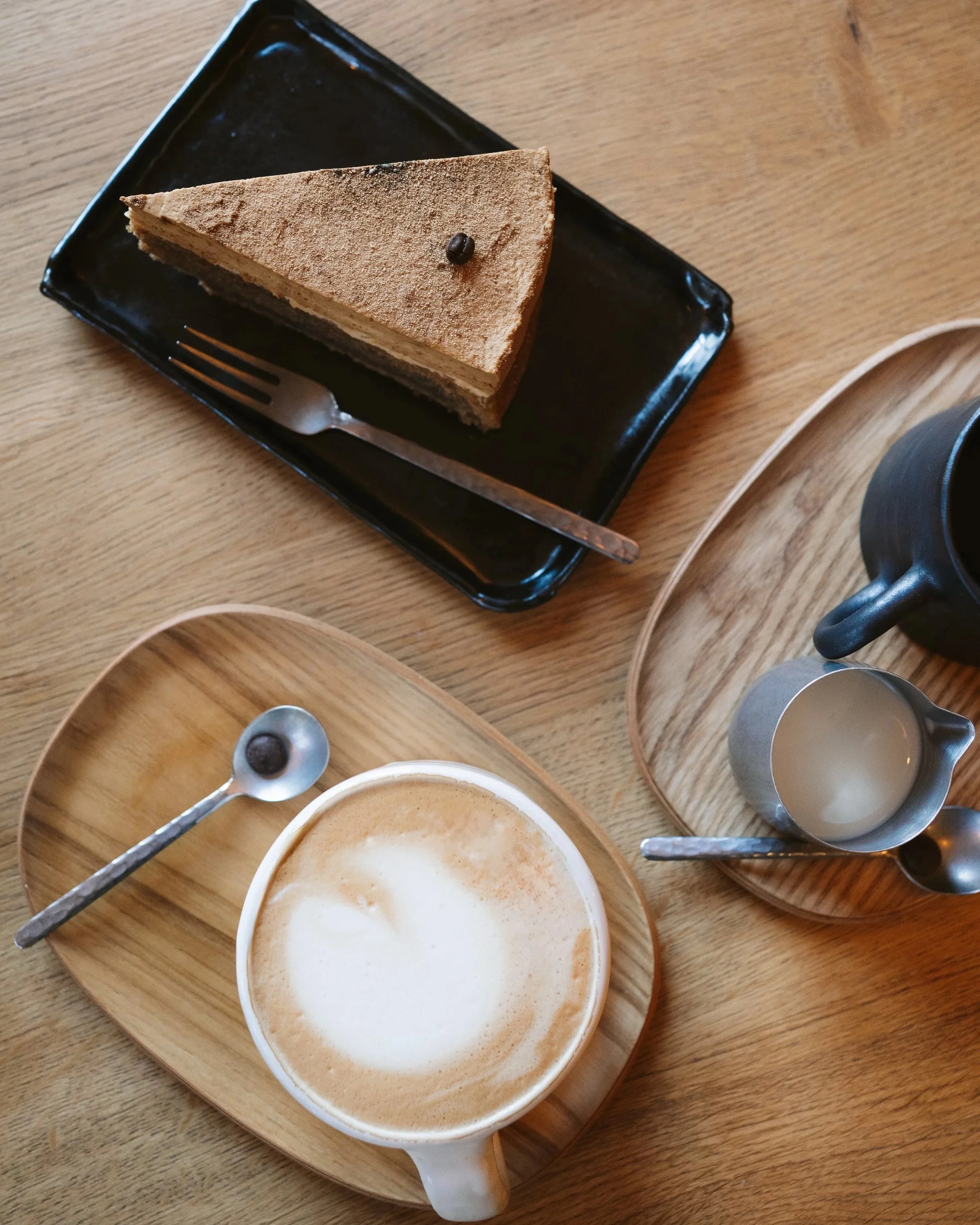 Ein Stück Tiramisu-Kuchen auf einem schwarzen Teller, ein Cappuccino mit Milchschaum auf einem Holztablett, eine kleine Kanne mit Milch und ein kleiner Löffel auf einem weiteren Holztablett