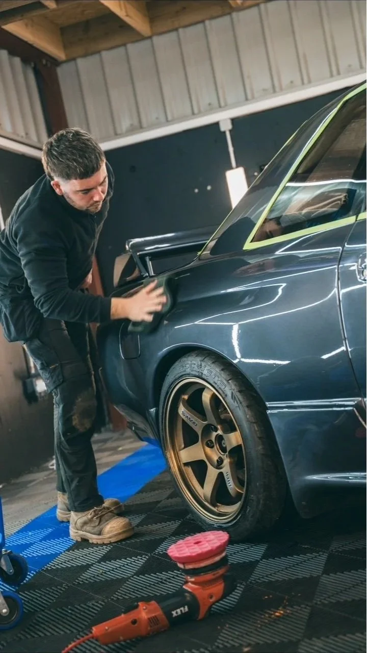 A man is polishing a black car inside a garage, with gold rims on the wheels, and a machine on the floor nearby.