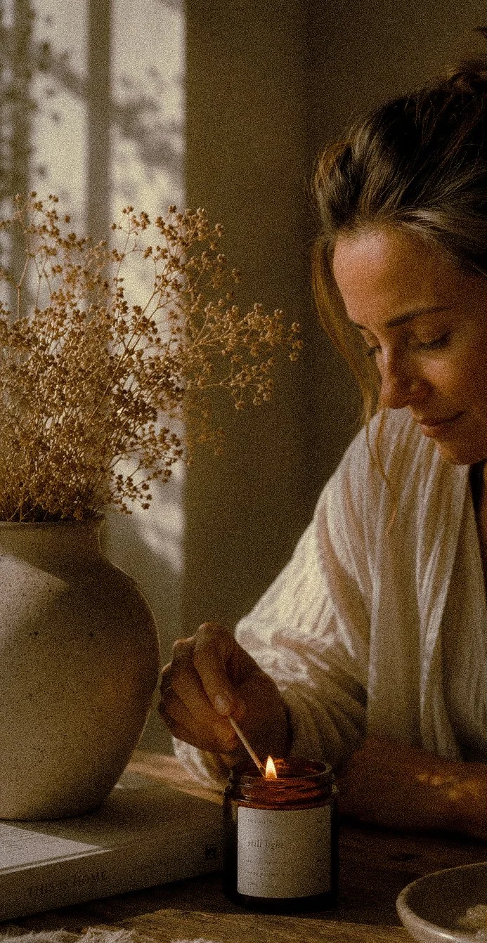 A woman lighting a aromatherapy candle with a matchstick on a table with a large vase of dried flowers nearby.
