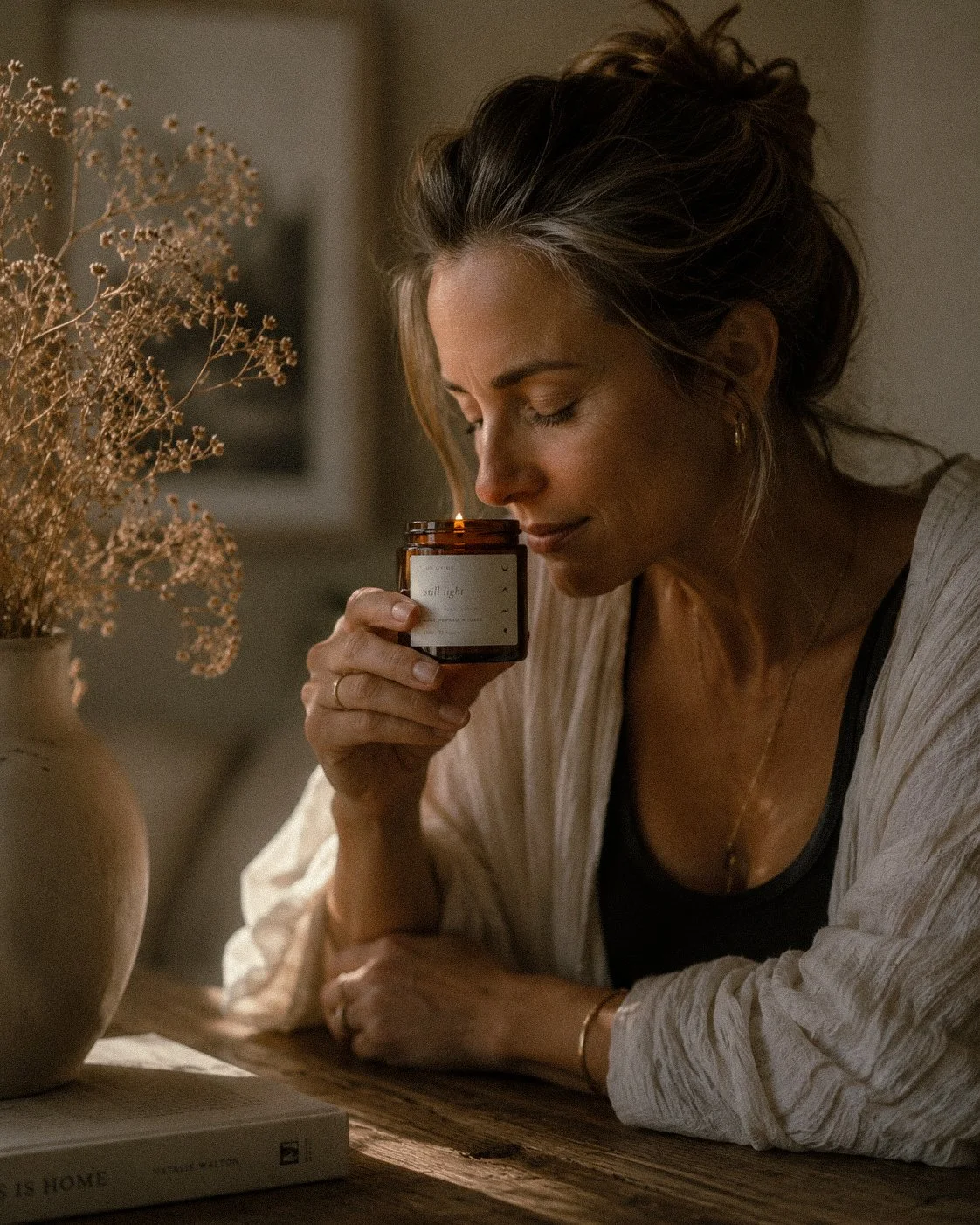 A woman with wavy hair, wearing a black top and a light cardigan, holds a lit natural scented candle close to her face while sitting at a wooden table. There is a vase with dried flowers and a book titled 'This Is Home' on the table.