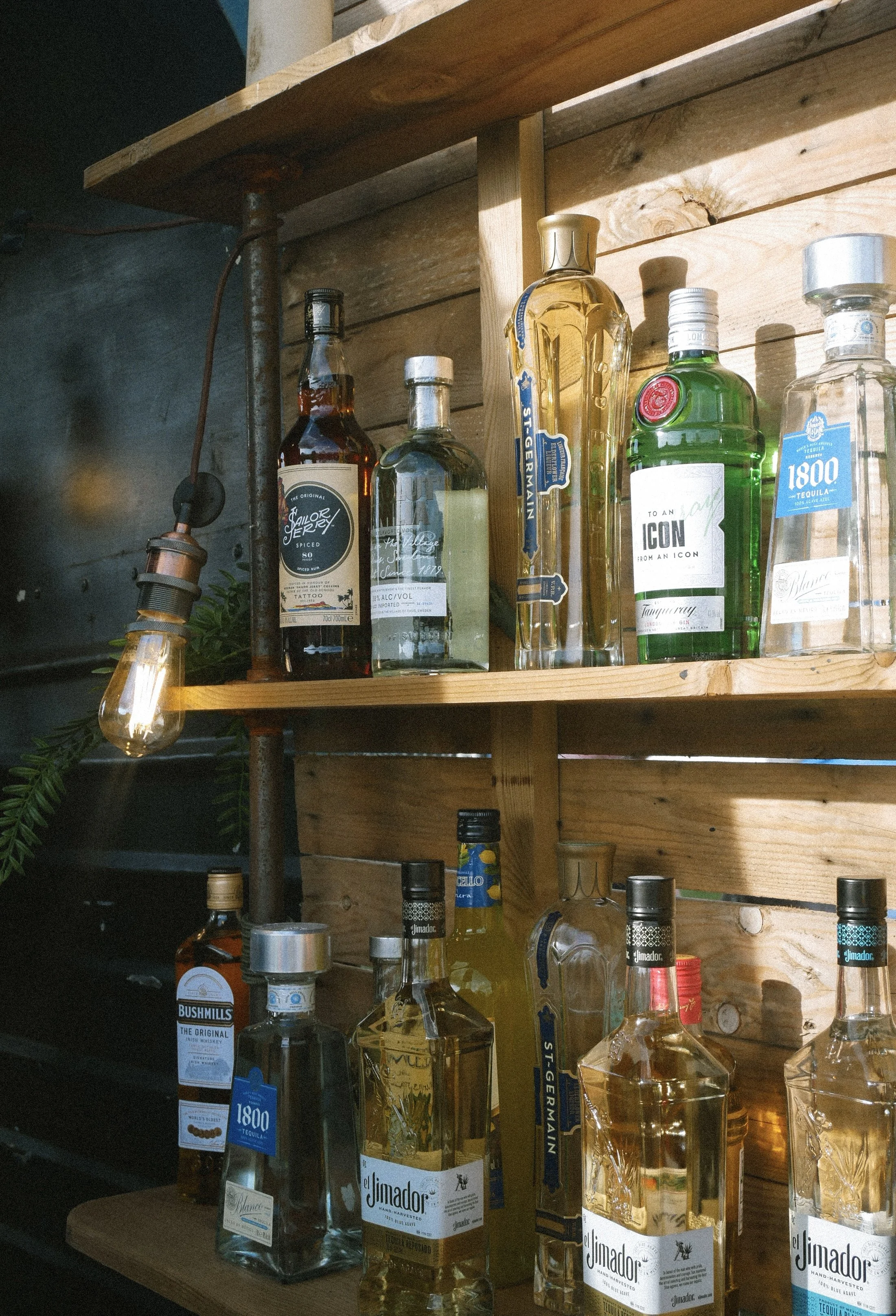 Arrangement of liquor bottles on wooden shelves in a bar or restaurant setting, with a lit hanging light bulb.