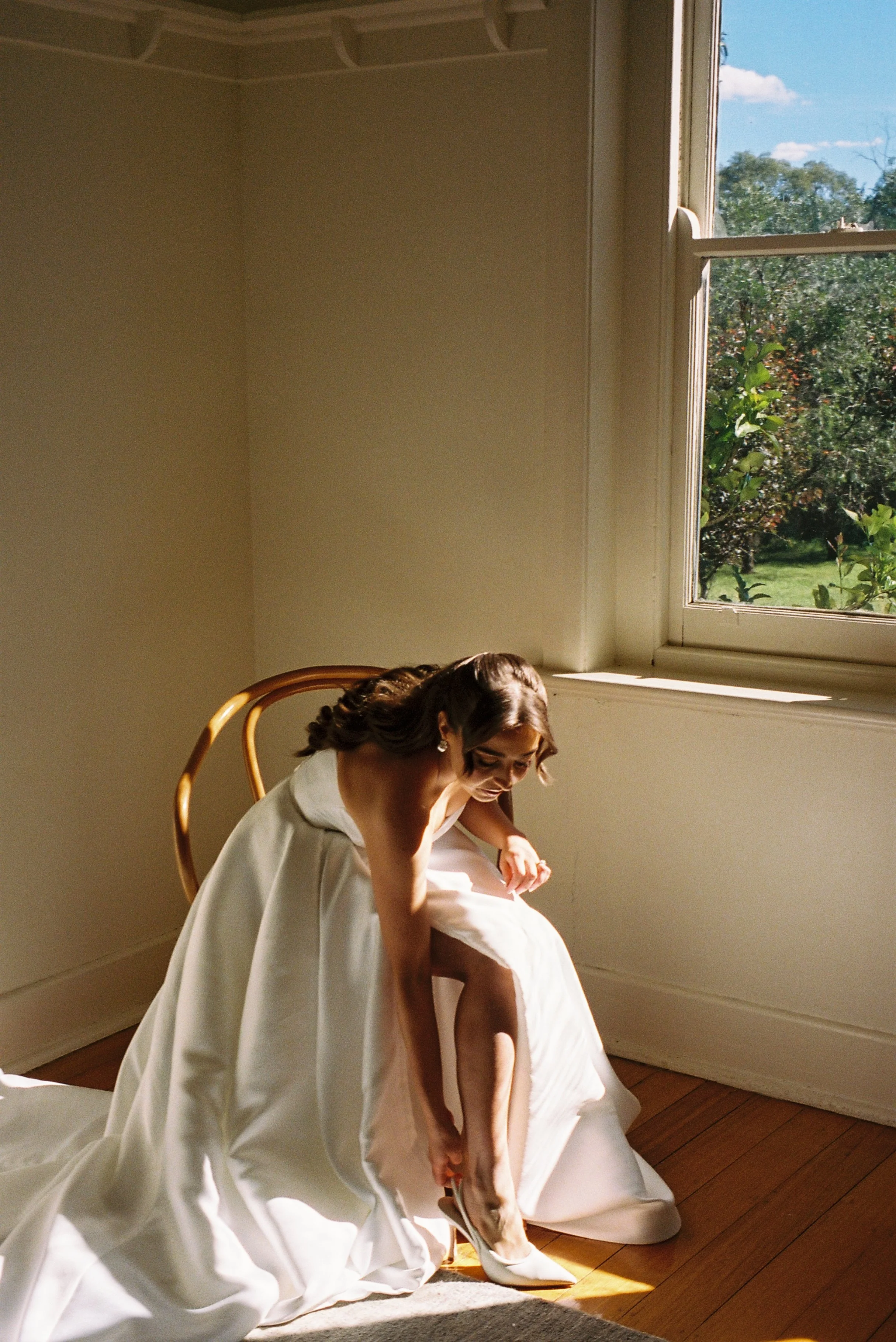 A woman in a white dress is sitting on the floor of a sunlit room near an open window, adjusting her shoe.