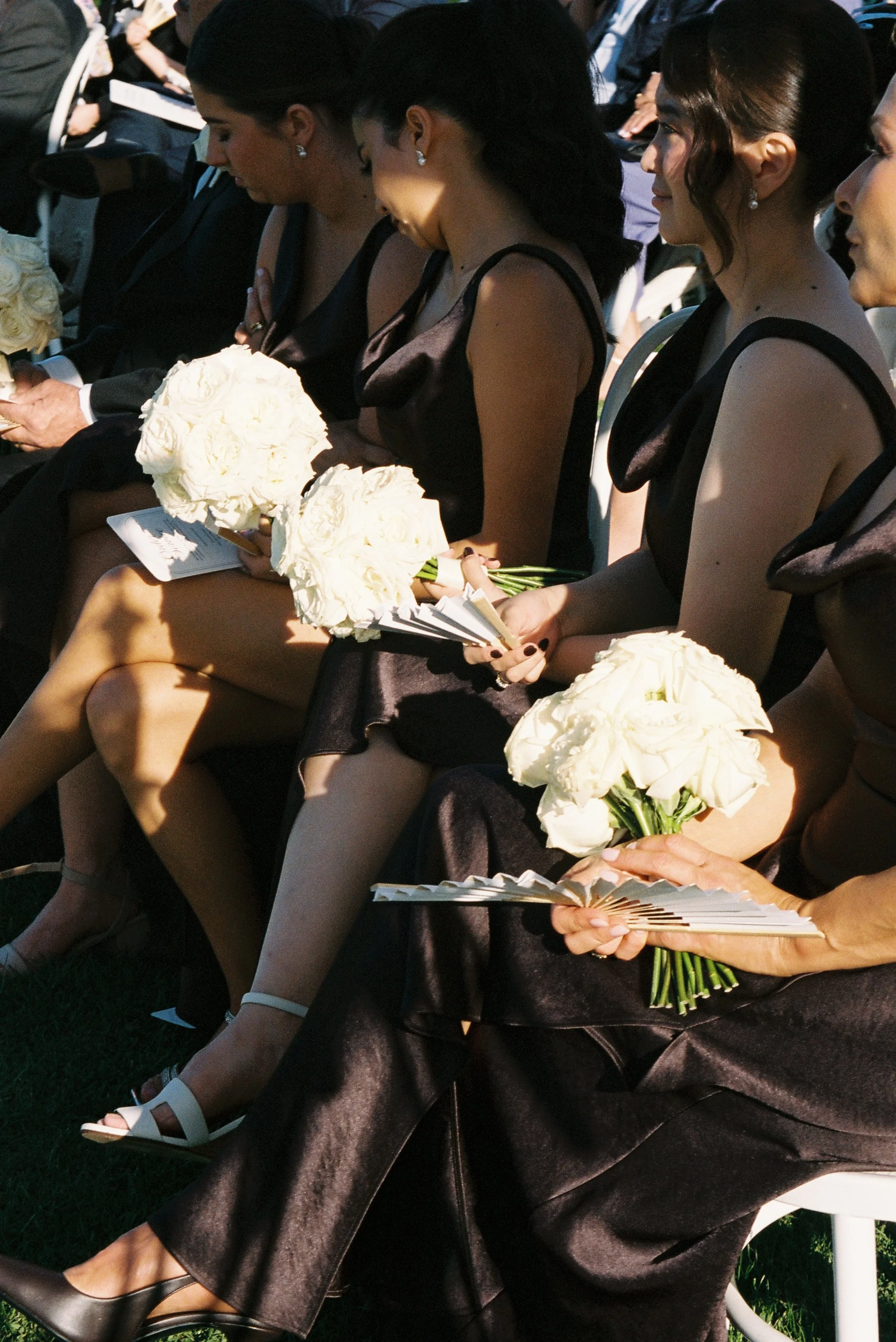 Women dressed in brown silk dresses with their hair styled elegantly, sitting in a line, holding white bouquets, at a wedding.