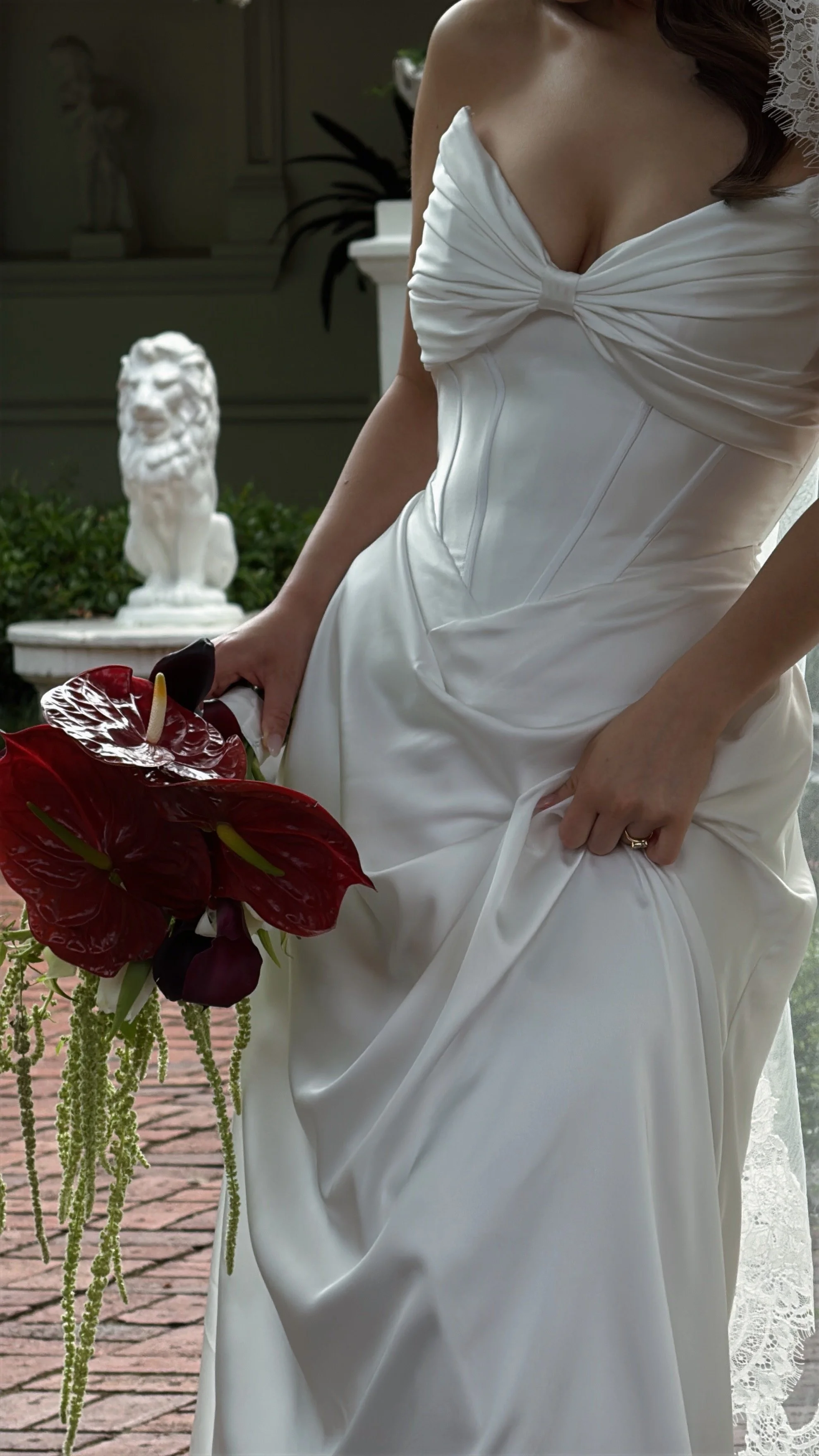 A woman in a white wedding dress holding a bouquet of red flowers, with a statue of a lion in the background.