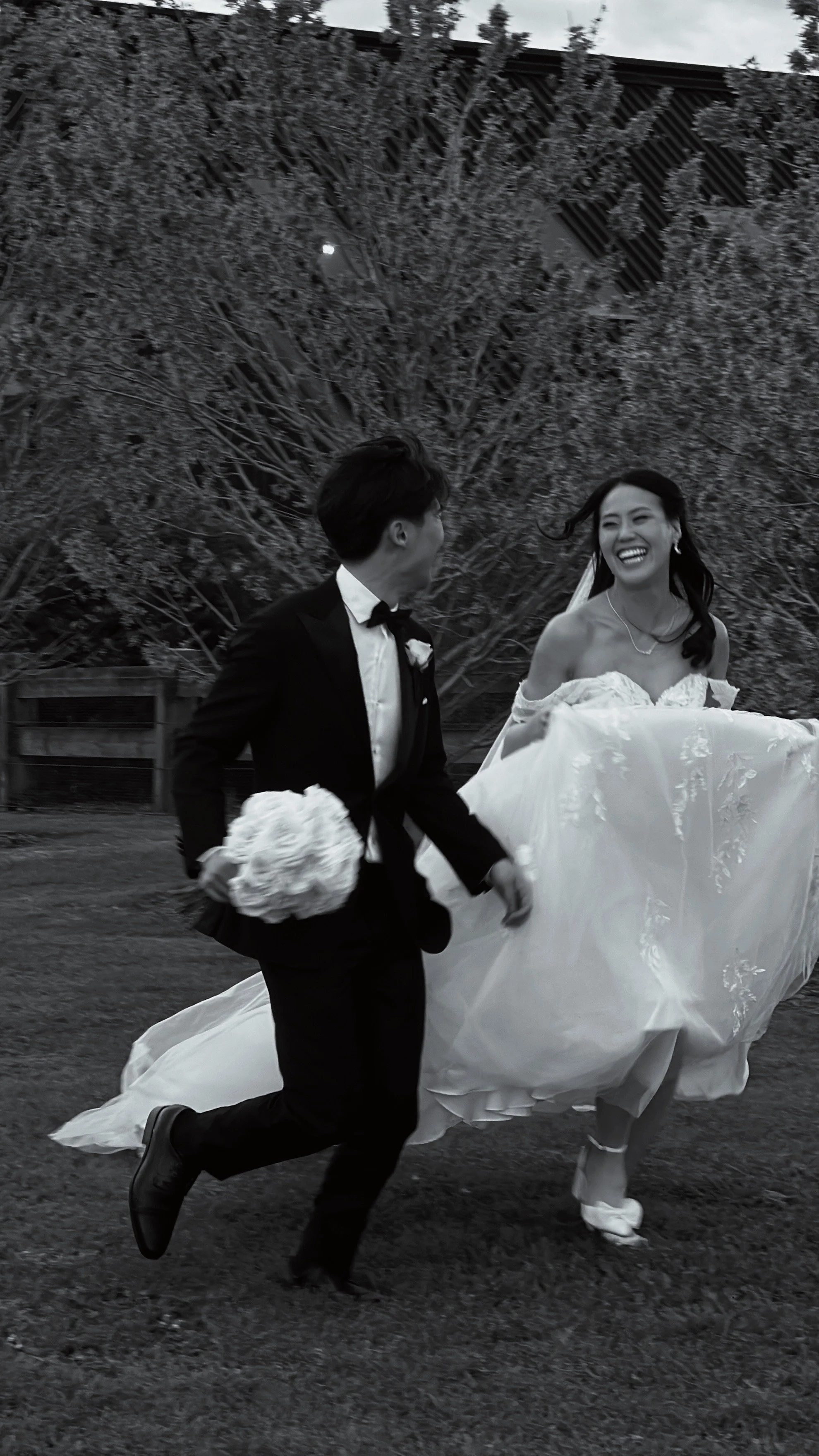 A black and white photo of a couple, the man in a tuxedo and the woman in a wedding dress, running outdoors while laughing and holding hands.
